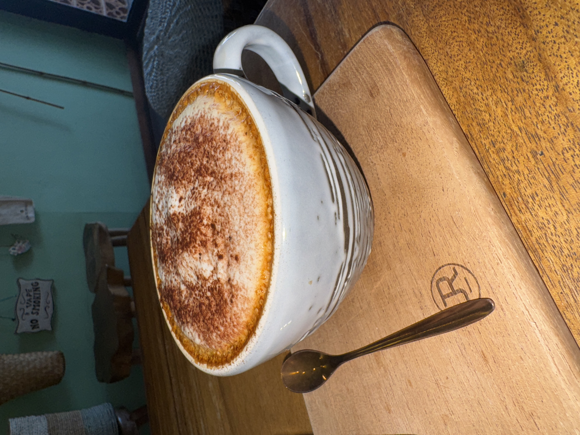 The image presents a close-up, overhead view of a coffee drink, arranged on a light brown wooden serving board. The central focus is a large, white ceramic mug, featuring a sturdy handle and a textured exterior with subtle vertical, light brown glazed streaks. The mug holds a foamy beverage, most likely a cappuccino or latte, generously topped with dark brown cocoa powder that covers most of the foam. A distinct golden-brown crema ring is visible around the inner edge of the cup.
To the right of the mug, a small, dark metallic spoon rests horizontally on the wooden board. The board itself exhibits a fine wood grain and has a faint, circular logo imprinted near the spoon, containing what appears to be a stylized letter "R."
The background, largely out of focus, reveals a teal-colored wall or surface. Partial glimpses of various decorative elements, possibly woven baskets or textured fabrics, can be seen. A small, rectangular sign is clearly visible on the wall, bearing the text "VAPE NO SMOKING." The overall scene suggests an indoor setting, likely a cafe or similar establishment, possibly located in Kuta Selatan, Indonesia, depicting a moment of enjoying a hot beverage.