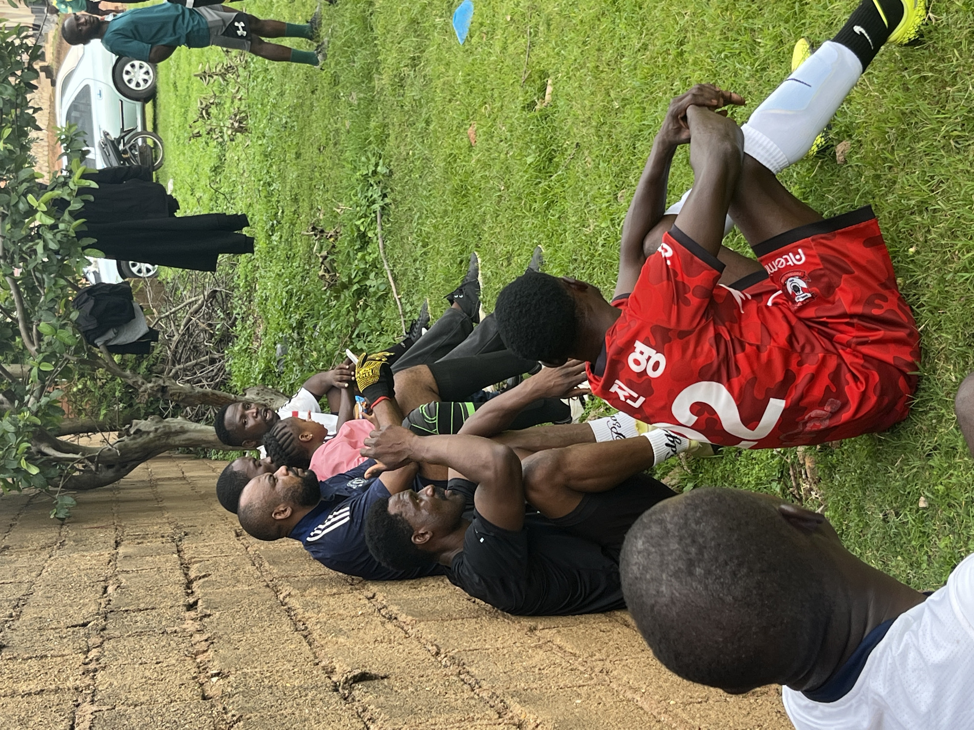 In this horizontal, eye-level outdoor shot, a group of young Black men, presumably a football team, are resting on a patch of ground with a brick-like texture. They are lying down, appearing to be in a post-training or pre-game session.
One individual in the foreground is wearing a red jersey with white numbers "2" and "80" visible, and black shorts. He has on white socks with black stripes and a white shin guard. Another person next to him is in a navy blue shirt with white stripes on the sleeves. Others are in dark clothing. The scene is set on a grassy area, with some trees and branches to the left. A car and a motorcycle are visible in the background, suggesting an informal training ground or park. The lighting indicates it is daytime, likely afternoon, with overcast or diffused sunlight. The overall mood seems relaxed and casual, with the individuals taking a break. No explicit text indicating the team name or sponsors is clearly legible, except for small logos on the red jersey. The location is identified as Jos, Nigeria.