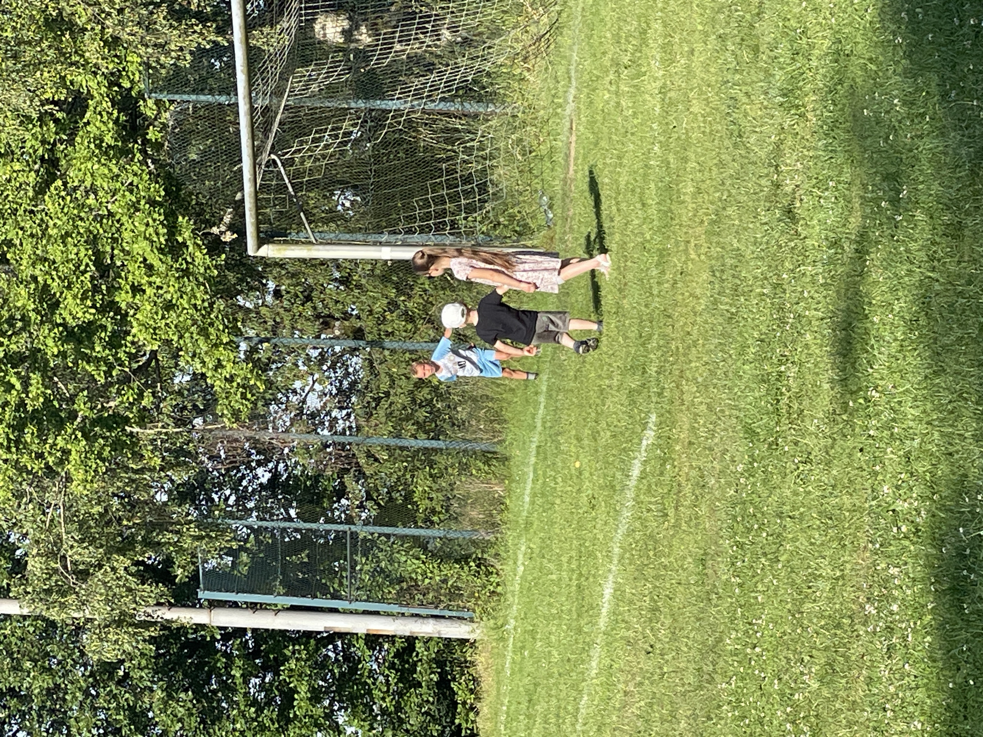 The image depicts a grassy field with a soccer goal in the background. Three children are visible on the field. A boy wearing a light blue shirt with the number "10" and dark shorts stands in the mid-ground. In the foreground, a boy in a black shirt and grey shorts, wearing a white cap, is walking away from the camera. To his right, a girl in a light-colored floral dress with long brown hair is also walking away from the camera. A soccer net is attached to the goalpost. Trees and a fence are visible behind the goal. The scene appears to be outdoors during daylight. The location is Beltershain, Germany.