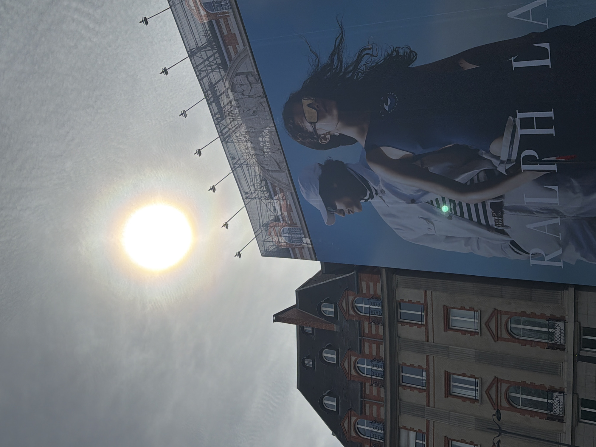 The image shows a bright sun in a cloudy sky next to a large billboard advertisement for Ralph Lauren. The billboard features two models. Below the billboard is a traditional Parisian building with a dark roof, arched windows, and red brick accents. The scene appears to be in Paris, France, due to the architectural style.