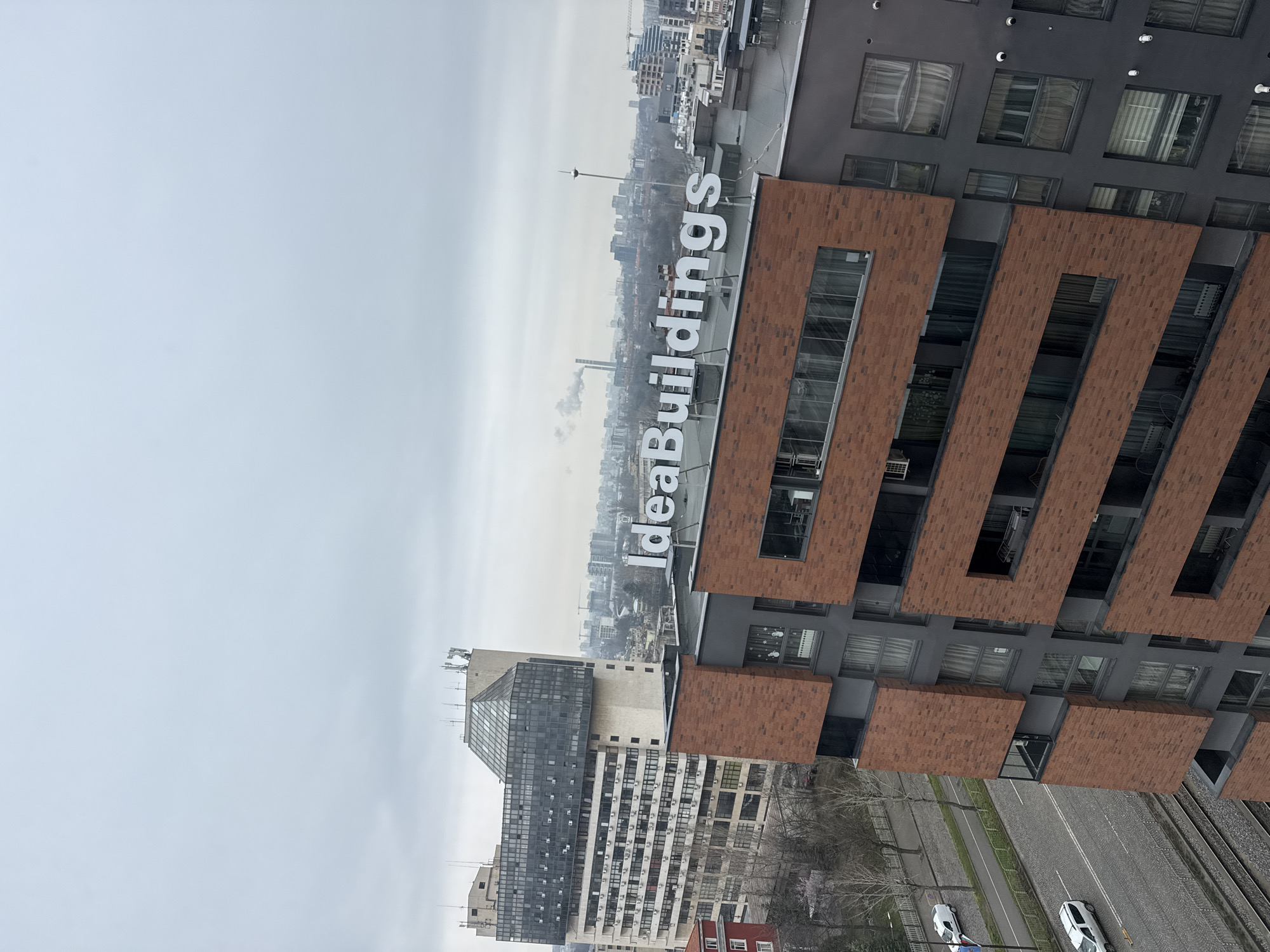 The image shows a high-angle view of a city landscape, identifiable as Sofia, Bulgaria, due to the "IdeaBuildings" signage on a modern building. The architectural style of the surrounding buildings includes a mix of old and new structures, typical for Sofia. The buildings display a range of materials including brick, concrete, and glass. The sky is overcast, suggesting a cloudy or possibly polluted day. On the street level, cars can be seen, indicating urban activity. There are no individuals visible in the image. In the background, a tall communications tower can be seen rising above the city skyline. In the background, a chimney with smoke.