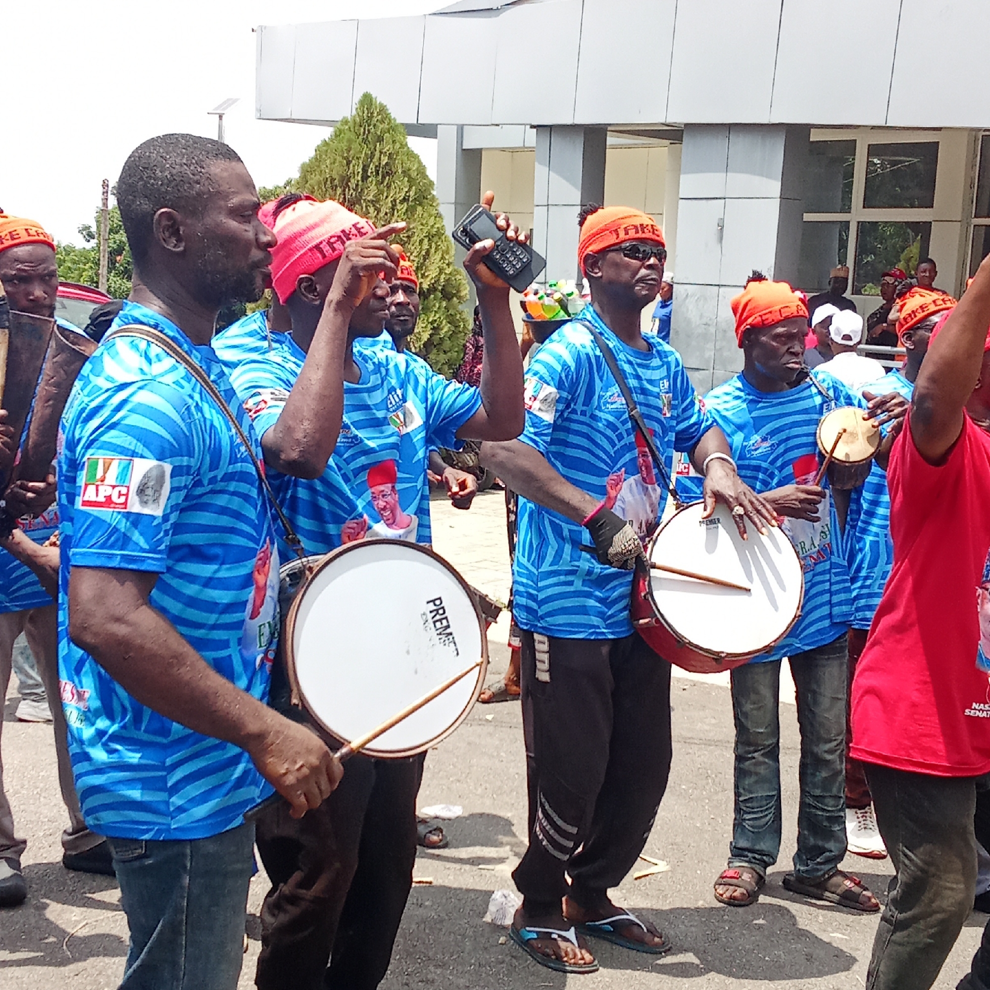 In Akwanga, Nigeria, a group of men dressed in blue shirts with blue and white stripes and orange knitted caps with the word "TAKE" marches in a procession. The shirts have APC logos on the sleeves and images of political figures on the front. Some men are drumming on drums strapped to their bodies; some hold smaller drums. One man is holding up a black cell phone. They are on a paved road, with a building in the background.