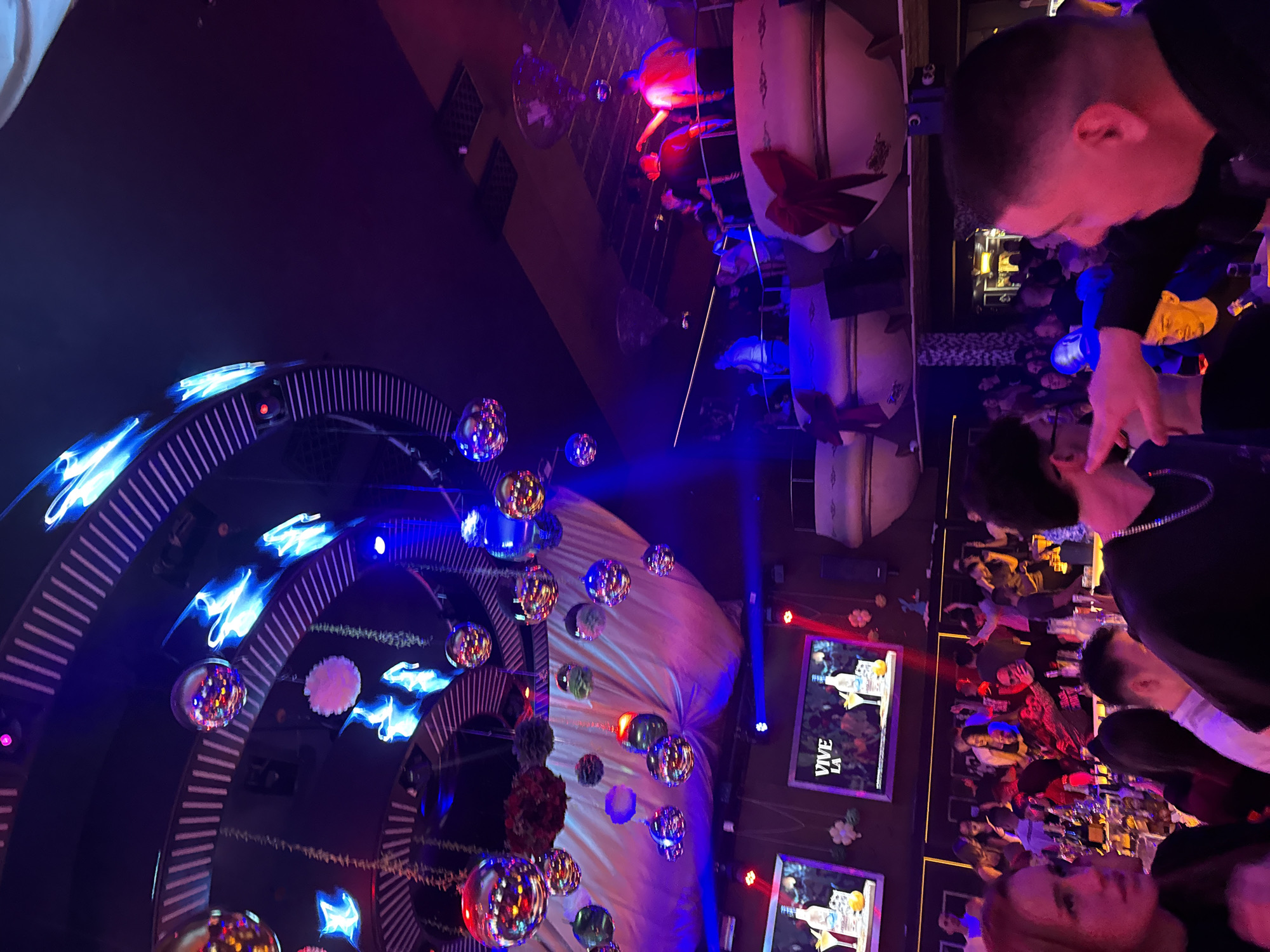 The image depicts a nightclub scene, likely in Pleven, Bulgaria. The foreground contains people, with one man predominantly featured on the right side of the frame, engaged in an unclear interaction. The background reveals a lively environment featuring multiple tables with patrons, illuminated by vivid lighting, and screens showcasing "VIVE LA" displayed on them. On the left side, there's an ornate circular structure decorated with blue lights and hanging disco balls. Above, one can see an elevated area with performances or activities, potentially a band setting the atmosphere. The overall ambiance is characteristic of a busy nightlife venue.