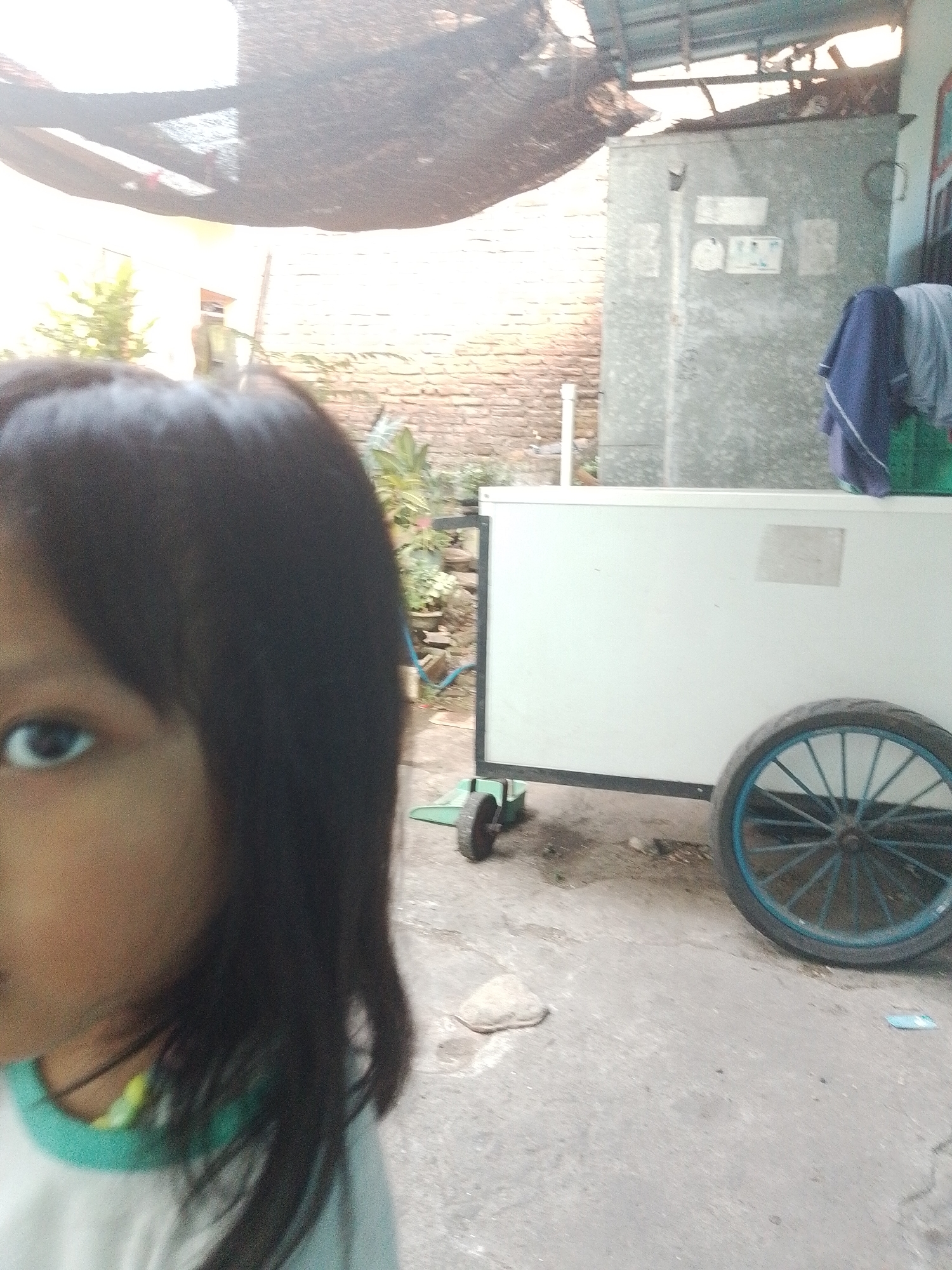 In an outdoor setting, likely within Kecamatan Pati, Indonesia, a close-up, waist-up shot focuses on the left side of a young girl's face and shoulder. Her dark hair is casually styled, falling around her face. She wears a light-colored shirt with a teal neckline.
In the background, a white, rectangular cart with a large spoked wheel (painted blue) on the right is visible. A smaller, dark wheel is present on the left side of the cart. To the left of the cart, a grey, rectangular structure, possibly a refrigerator or storage unit, stands against a brick wall. Clothes, including a blue garment and a gray one, are draped over the top of this structure.
Overhead, a dark, textured covering, possibly a tarp or awning, stretches across the frame, creating dappled light and shadows. The ground is paved with concrete. The overall impression is of a casual, everyday scene in a semi-urban or residential area.