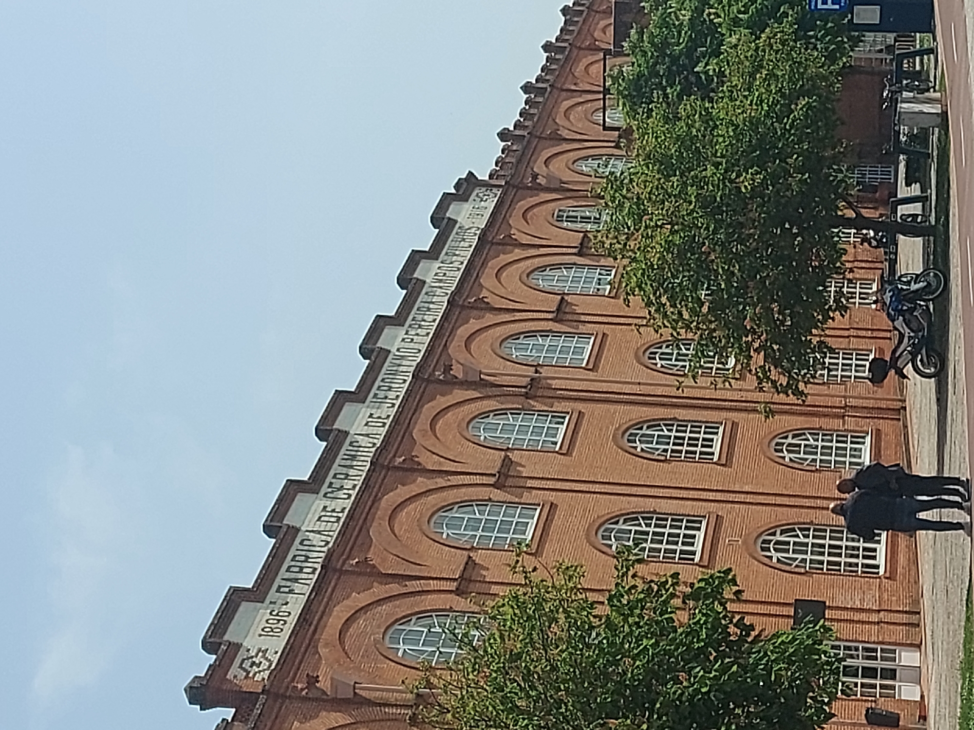 The image shows the Fábrica de Cerâmica Jerónimo Pereira Campos in Aveiro, Portugal. The brick building has rows of arched windows. Text on the building reads "1896 FABRICA DE CERAMICA DE JERONIMO PEREIRA CAMPOS FILHOS 1916-1958." Two people stand in front of the building. A motorcycle is parked nearby. Trees are in the foreground. The sky is visible in the upper left corner.