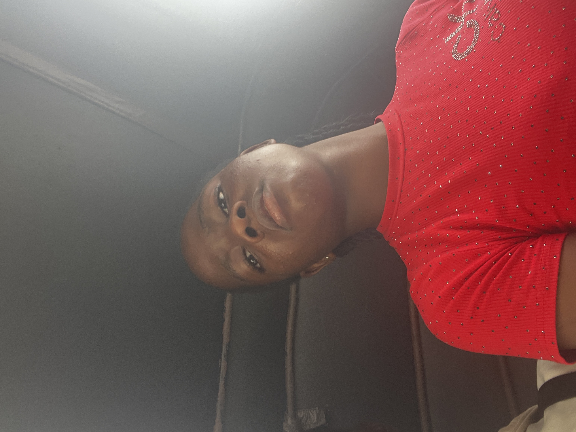 A young Black person with dark skin and braided hair is the subject of this portrait-style photograph. They are wearing a vibrant red, short-sleeved shirt adorned with numerous small, shimmering embellishments. The shirt also features a subtle graphic design made of similar sparkly material on the upper chest area, which appears to be letters or a symbol. The person's face is tilted, with their eyes looking slightly upwards and to the left. Their lips are parted, suggesting a gentle expression. The background is dark and somewhat indistinct, with a brighter area towards the upper left corner of the frame, possibly indicating a light source or a window. The overall lighting suggests an indoor setting, with the light catching the person's face and the details of their shirt. The orientation of the image is such that the person's head is to the left, and their body extends towards the right and downwards. The image quality is somewhat grainy, which is common for photographs taken in lower light conditions. The context provided is Nkpor, Nigeria, though no specific location cues are evident within the image itself.