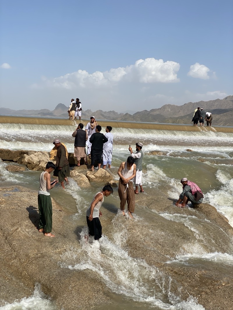 The image shows a group of people at a waterfall in what appears to be a rocky, arid environment. Some people are standing on the edge of the waterfall, while others are in the water. Most are wearing traditional Afghan clothing. A few are using phones to take pictures. The waterfall is flowing over a man-made structure, likely a dam or weir. In the background, there are mountains. There is no indication the location is in the city of Kim, Afghanistan.