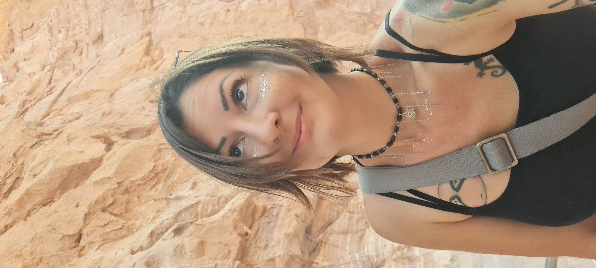 A woman with short brown hair is smiling and looking at the camera. She has glitter under her eyes and a nose ring. She is wearing a black tank top and a black choker necklace. A gray strap is slung across her body. She has tattoos on her arms and chest. The background appears to be a textured rock wall.
The location cannot be pinpointed more specifically within Hevel Eilot Regional Council, Israel, based on the image alone. Further context is needed.