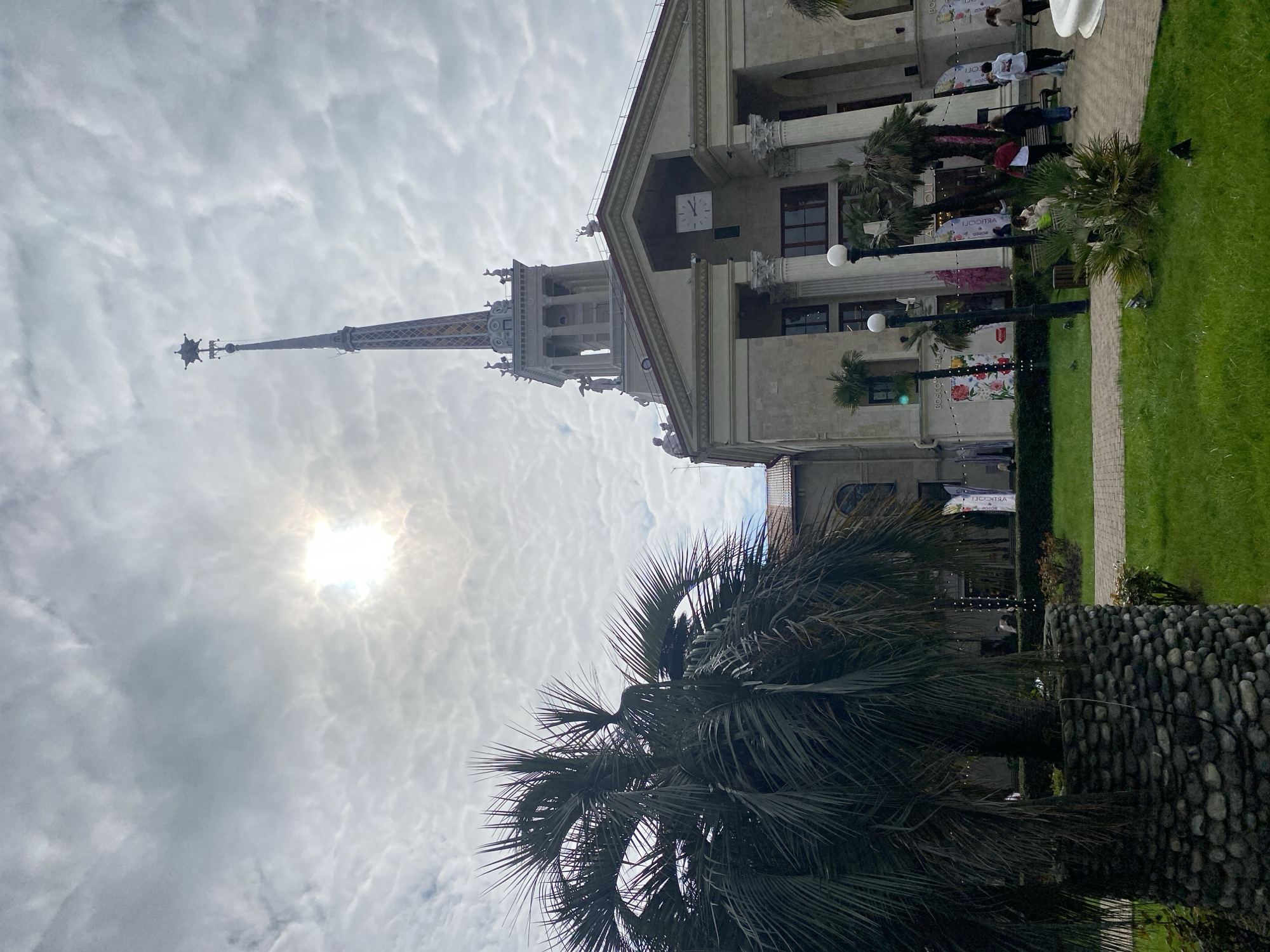 The image displays the Sochi Seaport building (Marine Station) in Sochi, Russia, under a cloudy sky with the sun faintly visible through the overcast conditions. The prominent structure is a multi-tiered building featuring Stalinist Empire style architecture, characterized by a tall central spire topped with a star, classical columns, and numerous statues adorning the roofline and tower levels. A large square clock face is visible on the upper facade of the main building section.
In the immediate foreground, to the left, a large palm tree with dark, spiky fronds is prominent, its trunk encased in a cylindrical planter made of stacked grey stones. To the right of the palm tree and extending towards the building, a manicured green lawn is visible, traversed by paved pathways. Several spherical white lampposts are spaced along these pathways.
Multiple individuals are observed on the paved areas near the building, engaged in casual pedestrian activity; some are walking, while others appear to be seated on benches. They are dressed in various casual attire. Near the building's entrance, several colorful vertical banners featuring floral and abstract patterns are displayed. Additional palm trees are situated closer to the building's facade. The scene depicts a public urban environment in front of a significant architectural landmark.