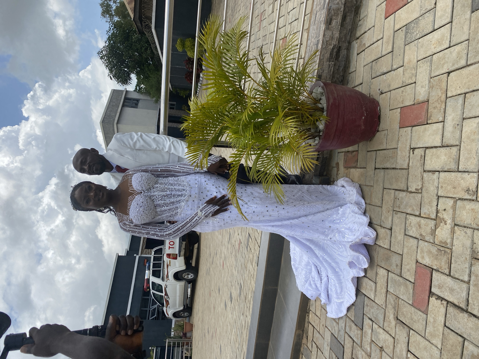 A full shot captures a wedding couple standing outdoors on a paved patio in Bukuru, Nigeria. The bride, a Black woman, wears an elaborate, floor-length white wedding dress adorned with sparkling beads and sequins. The dress features a sweetheart neckline, long sleeves, and a mermaid silhouette that flares out at the bottom. She has dark, curly hair and is wearing earrings and a tiara. The groom, a Black man, stands beside her, dressed in a white suit with a red tie. He has a bald head and a goatee.
Behind the couple, a white Toyota pickup truck with flashing red and blue lights is parked. The truck has "TOYOTA" printed on the tailgate. In the foreground, a large potted plant with green, palm-like leaves sits to the right of the couple. The environment includes a building with dark siding and large windows in the background, as well as a metal railing. The sky is cloudy, with patches of blue visible. The ground is paved with interlocking bricks of various beige and red tones. A hand holding a professional video camera is visible on the left side of the frame, suggesting a wedding videographer is present.