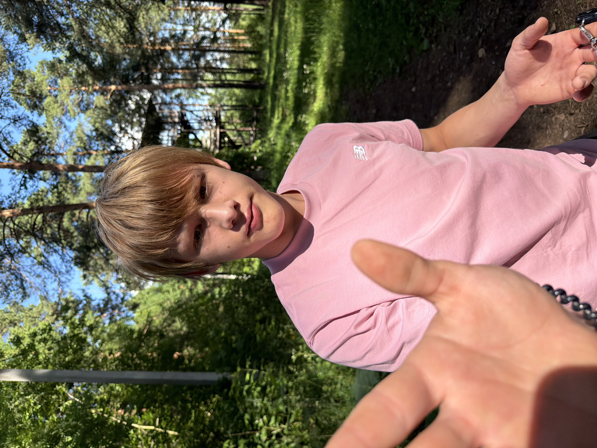 A young person with light brown hair and fair skin is standing outdoors, looking at the camera. They are wearing a pink t-shirt with a white logo on the left chest. Their left hand is extended towards the camera, slightly out of focus. Their right hand is open, palm up, and positioned to the right of their body. The background consists of a forest with tall pine trees and green foliage. Sunlight filters through the trees, creating dappled lighting on the ground. In the distance, a wooden structure with platforms and stairs is visible, suggesting a rope course or adventure park. The location is identified as Izhevsk, Russia.