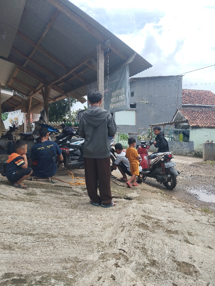 The scene takes place in the city of Kutawaringin, Indonesia, at latitude -6.974 and longitude 107.519. A group of five men are gathered under a corrugated metal roof overhang in front of a building with a faded sign. Several motorcycles are parked in the area. One man is crouched down, seemingly fixing one of the motorcycles, while another stands by the side of the bike. There is a yellow air hose running from the ground towards the bike, and a small yellow air compressor nearby. Two men are standing in the center of the scene, facing the camera, looking at the men working on the motorbike. A young child in yellow clothing stands in front of the motorbike, looking at the men working on it. The ground is covered in a layer of dirt and dust. The sky is cloudy and gray. The man on the right is wearing a blue shirt and black pants, while the other man is wearing a dark gray hoodie. The man crouching down is wearing a blue t-shirt and black pants, and the two men standing are wearing similar attire. The child is wearing a yellow shirt and blue shorts. There is a yellow air hose connected to the front tire of the motorcycle. The man fixing the motorcycle is bending over, with his back to the camera. The man standing next to the bike is facing the camera, but his face is not visible.