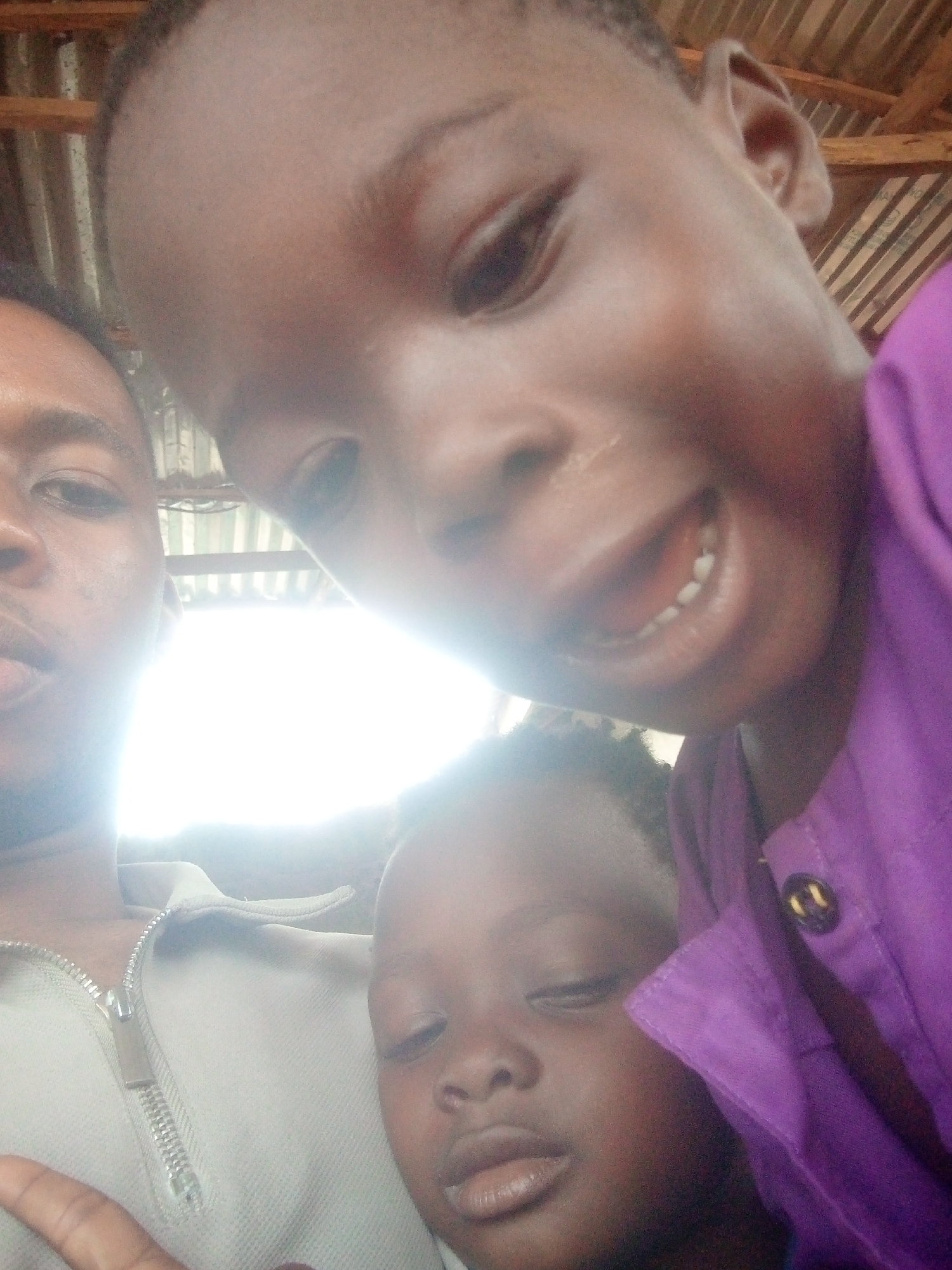 A selfie shows three individuals. An adult is on the left wearing a collared shirt with a zipper. Two children are positioned to the right of the adult, one in a purple shirt. The location appears to be indoors, with a corrugated metal roof visible overhead. The city and exact location are not discernible.
