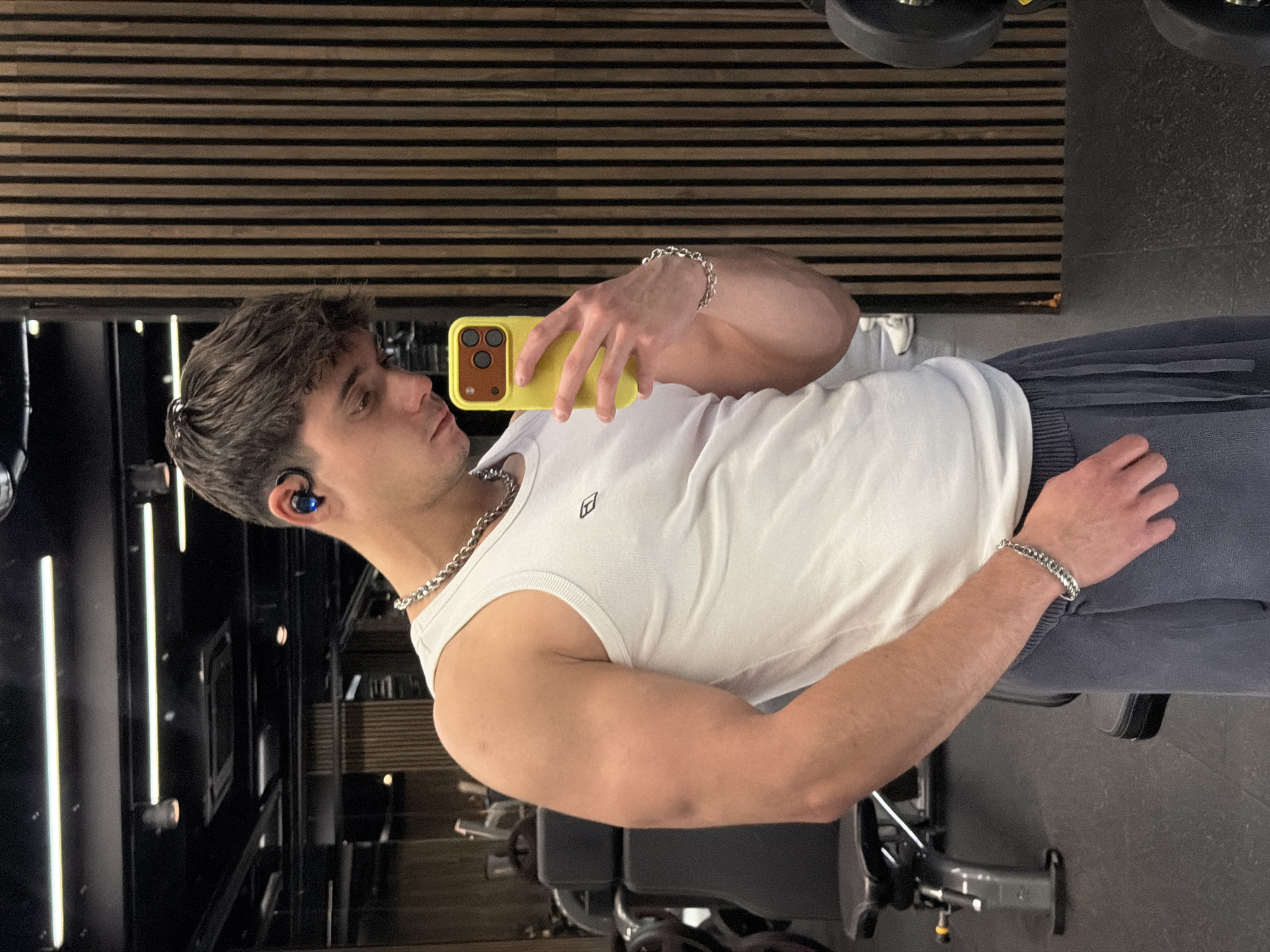 A young man poses for a photograph in a gym-like setting. He is wearing a white tank top and gray sweatpants. The subject is holding a yellow iPhone in his left hand, angled to capture his reflection in the mirror behind him. He has dark hair and an earbud in his left ear, adorned with a silver chain necklace and bracelet. He appears to be sitting on gym equipment. The background is a mix of wood paneling and gym equipment, with some lights and a textured floor. The specific location within Sofia, Bulgaria, cannot be determined from this single image.