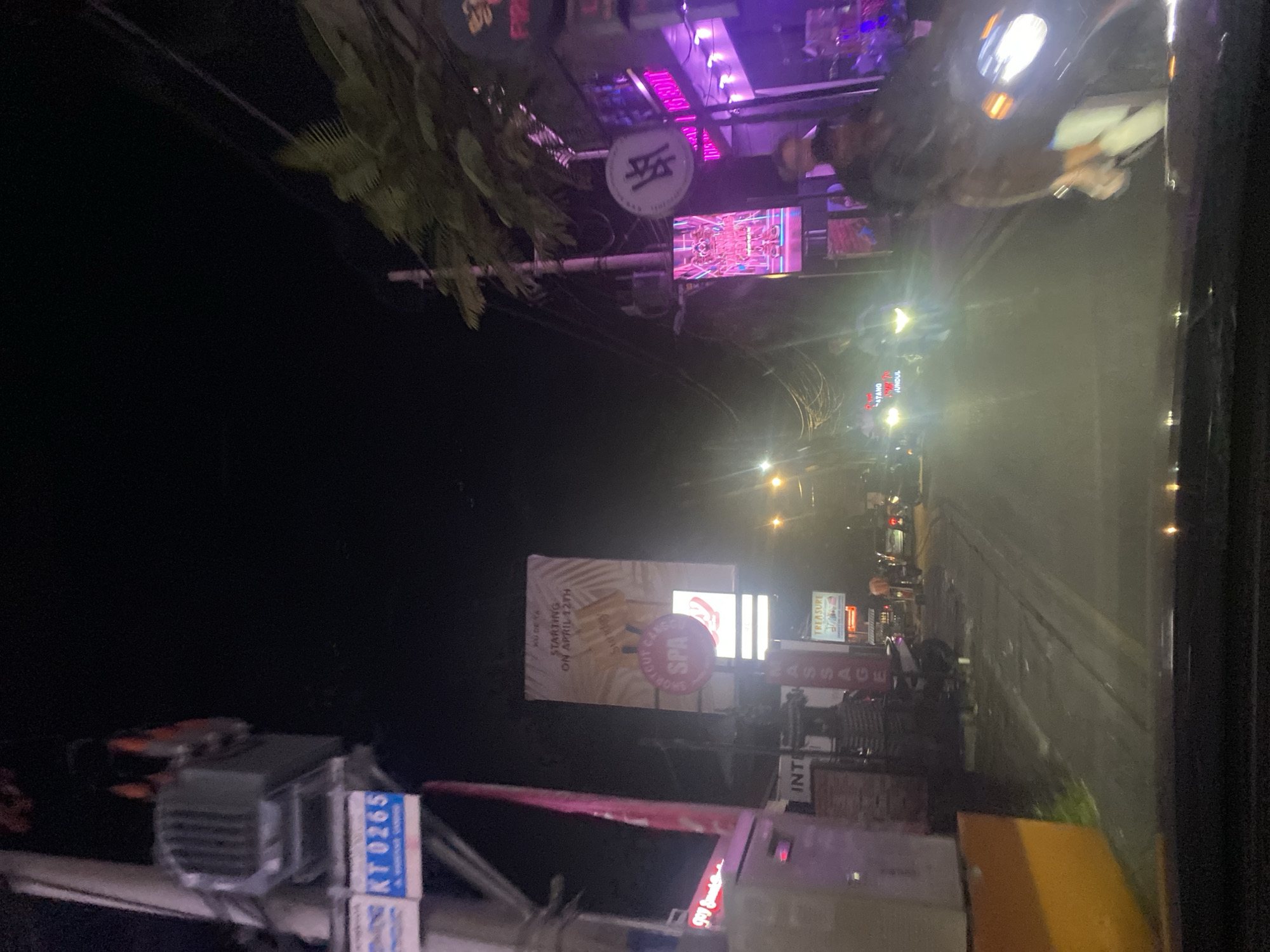 The image shows a nighttime view from a vehicle driving along a street in North Kuta, Indonesia. The street is lined with shops, restaurants, and businesses, illuminated by bright neon signs. Various signs advertise spas, massages, and restaurants, including one that says "Ku De Ta." The license plate "KT 0265" is visible on a nearby utility box. Some people are walking along the sidewalk and traffic is visible on the road.