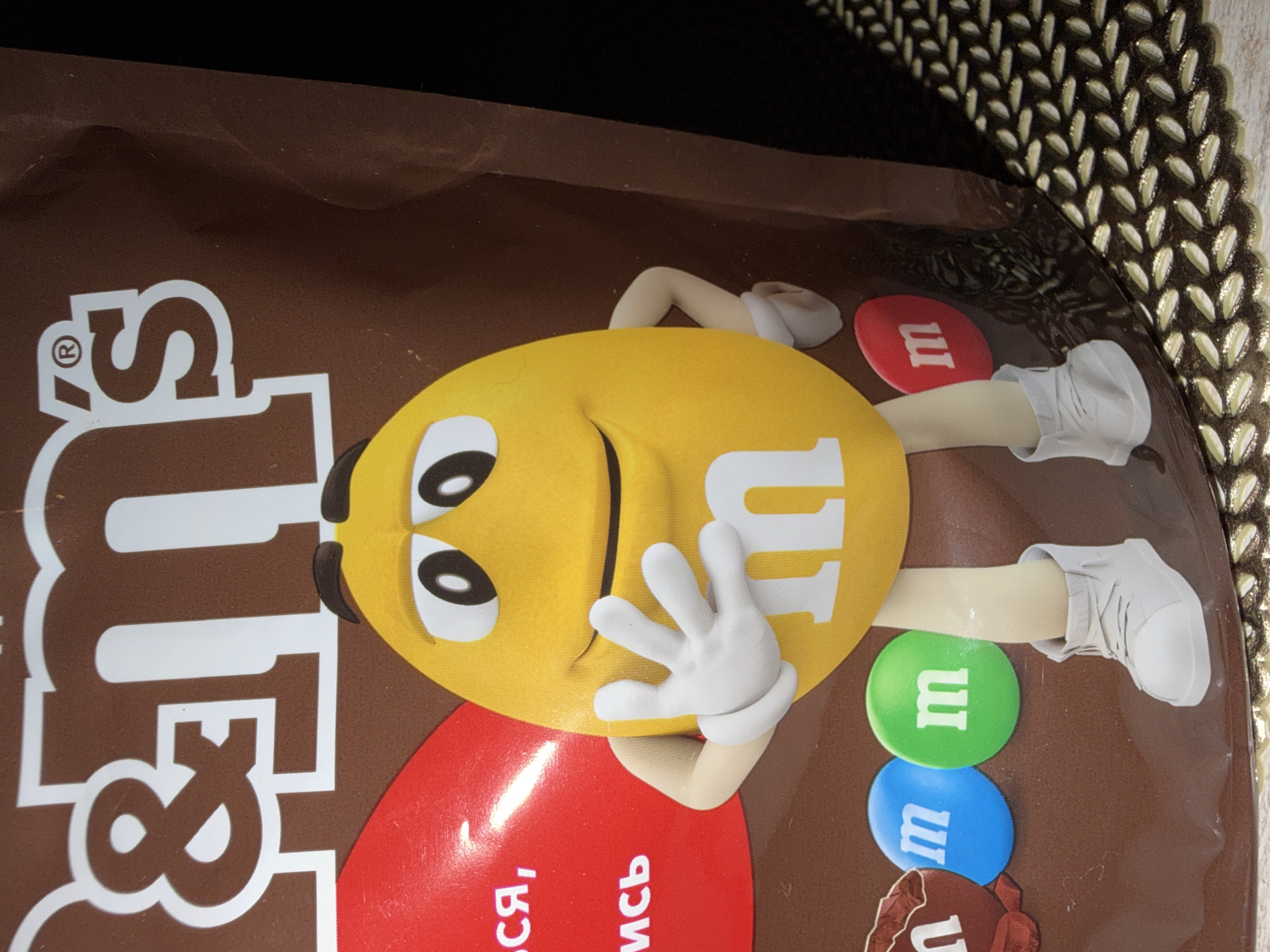 The image shows a close-up of a M&M's candy bag with the characteristic "m&m's" logo prominently displayed in white lettering on a brown background. In the foreground, a yellow M&M character with a smiling face and white gloves is featured. The character's left hand is raised, and its body is partially obscured by the bag's packaging. The character is wearing white sneakers. Below the character, three individual M&M's candies are visible: one brown, one blue, and one green, each with a white "m" on them. The background is dark, with a textured surface, possibly a placemat, visible on the right side of the image. There is no information in the image to suggest a location in Sirius, Russia.
