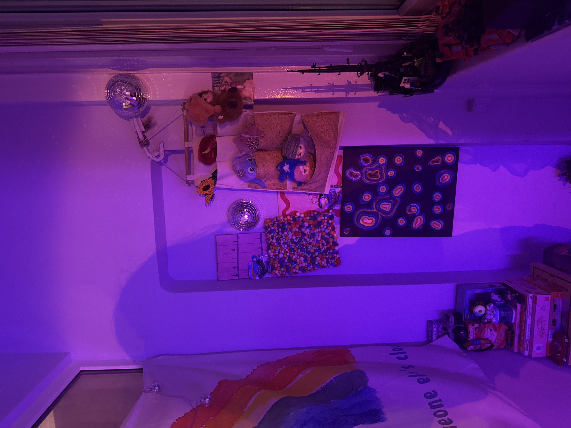 The image shows a bedroom interior with a purple light cast. The room features a bed with a rainbow-colored blanket and text on the pillow, reading "Someone el's clo". The wall is decorated with various items, including a disco ball, stuffed animals in a cork enclosure, a sunflower image, a round sequined object, a small dollhouse, a square of multi-colored balls, and a black painting with abstract shapes. Above, closer to the ceiling, are toy guns and Lego-like construction sets. Stacked on a table or surface to the right are books, boxes, and toys. There are blinds at the top of the frame. The exact location of Yerevan, Armenia cannot be determined from the image.