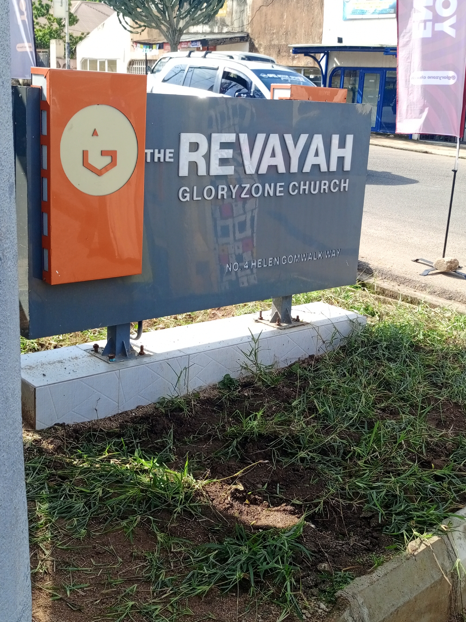 An outdoor, eye-level, medium shot captures a sign for "THE REVAYAH GLORYZONE CHURCH." The sign is a dark gray rectangular structure with white text, and an orange panel on the left side featuring a white circular emblem with an orange geometric design inside. Below the church name, the address "NO. 4 HELEN GOMWALK WAY" is also visible in white text. The sign is supported by dark metal poles bolted to a white tiled concrete base.
The area in front of the sign consists of a patch of dry soil with sparse green grass. A concrete curb borders the right side of the frame, with more grass and soil beyond it. In the background, a white building with blue trim and awnings is visible, along with parked cars. To the right of the church sign, a light purple flag on a black pole stands in the foreground. The overall scene suggests a street view in a commercial or residential area. The location is identified as Jos, Nigeria.