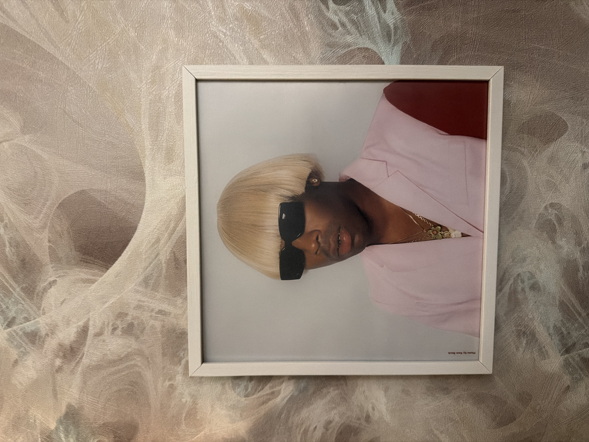 A framed photograph of Tyler, the Creator is displayed on a wall. The photograph features him with a blonde bowl cut wig and black sunglasses, wearing a light pink blazer with a dark red sleeve. He is also wearing a gold chain and grillz. The photograph is mounted on a textured wall with a swirled pattern in shades of beige and grey. The bottom left corner of the photograph shows the text "Photo by Ren Rick". The overall scene appears to be an indoor setting.