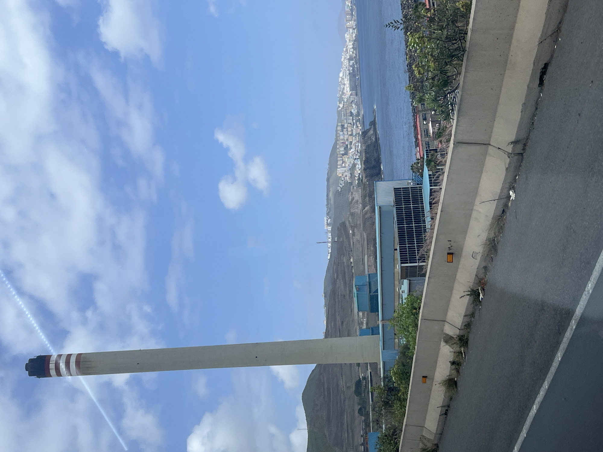 The image depicts a view from an elevated position, likely a road or bridge, overlooking the city of Las Palmas in Spain. The sky is mostly blue with scattered white clouds. A tall, cylindrical chimney with red and white stripes at the top dominates the lower left of the frame. Adjacent to the chimney is a blue industrial complex.
In the distance, a coastline stretches along the frame, where the city of Las Palmas is situated. The cityscape comprises many white buildings clustered together, descending towards the ocean. The ocean itself occupies the upper-right portion of the photo.
In the foreground, a gray concrete barrier runs along the right side of the frame, separating the viewer from the landscape. Some vegetation grows along the edge of the barrier. A road is visible next to the barrier.