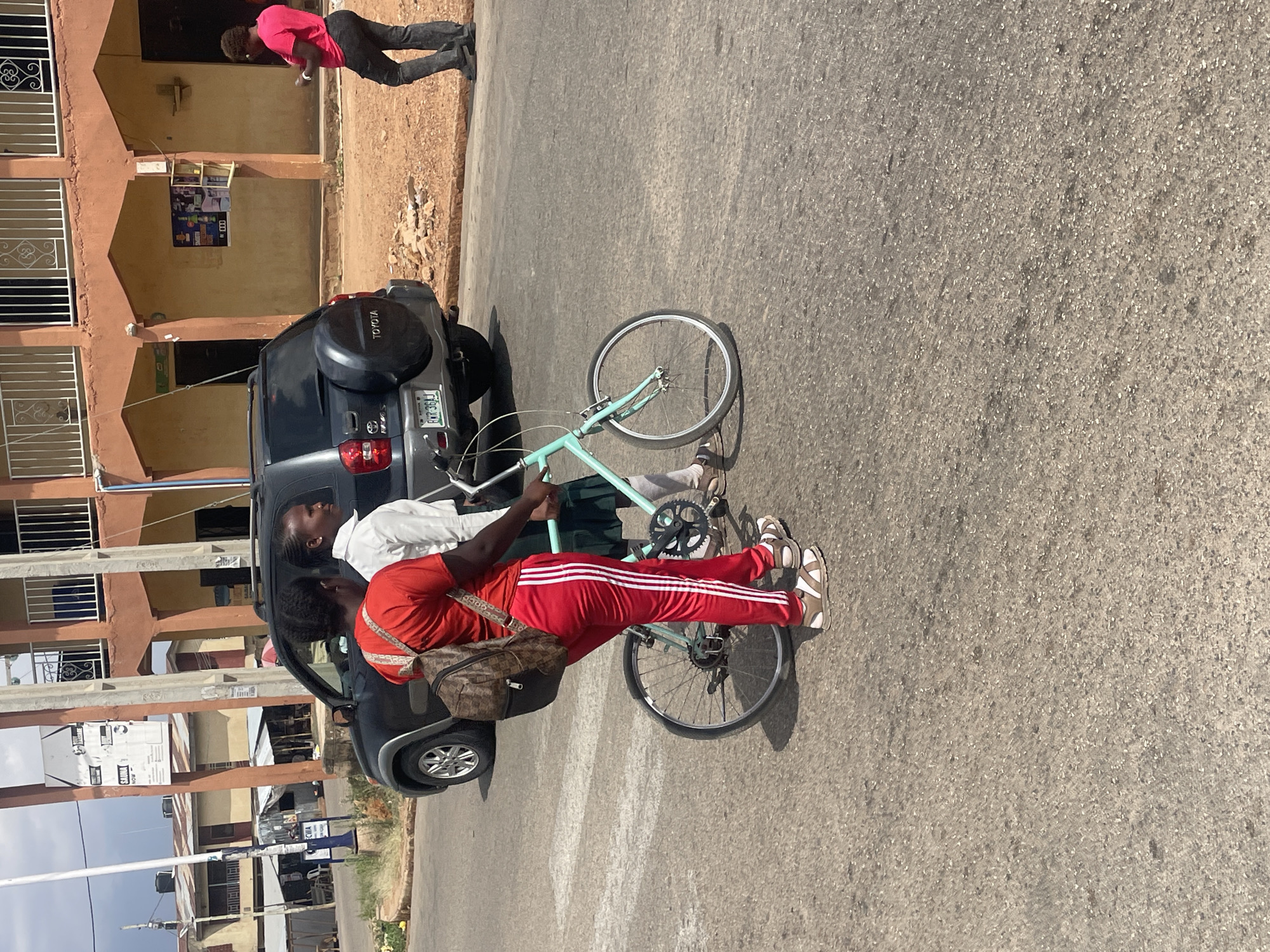 The image shows two individuals beside a dark-colored Toyota SUV with a covered spare tire mounted on the rear. The vehicle's license plate is partially visible, reading "RRR 361J". A light blue bicycle is positioned between the two individuals and the car. One individual is wearing a red shirt and red track pants with white stripes, along with brown sandals. The other is wearing a white shirt.
The scene appears to be a street in Jos, Nigeria. The background shows a multi-story building with brown exterior walls and white window frames. A pedestrian in a pink shirt and dark pants is walking in the background, next to the building. The street is paved with gray asphalt, and there are other buildings and signs visible further down the street.