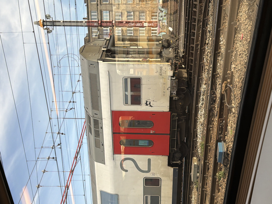 The image presents a detailed view of railway infrastructure and an adjacent urban environment, observed from an interior perspective through a window, located in Saint-Gilles - Sint-Gillis, Belgium.
A light grey and white double-decker railway carriage occupies the midground, positioned on one of several parallel tracks. The carriage displays a large grey numeral "2" on its left side and the identification number "62066" on a white panel below a smaller window to the right of the doors. Two prominent red double-doors with reflective windows are centrally located on the carriage's visible side. The lower section of the carriage, particularly around the doors and wheels, exhibits visible dirt and grime. The right end of the carriage shows coupling mechanisms.
The foreground and midground are dominated by multiple parallel railway tracks, constructed with steel rails resting on a bed of ballast. Various trackside equipment, including a blue rectangular box and a grey square box, are visible between the tracks. Overhead, a dense network of electrical catenary lines and support structures spans the upper portion of the scene, set against a predominantly clear blue sky with faint wisps of clouds.
In the background, a tall, red and white striped utility or signal mast, equipped with lighting elements at its apex, rises above the tracks. Behind this mast and extending across the horizon are several multi-story urban buildings featuring numerous windows. Graffiti is discernible on a lower wall or barrier situated in front of these background structures. No individuals are present in the depicted scene.