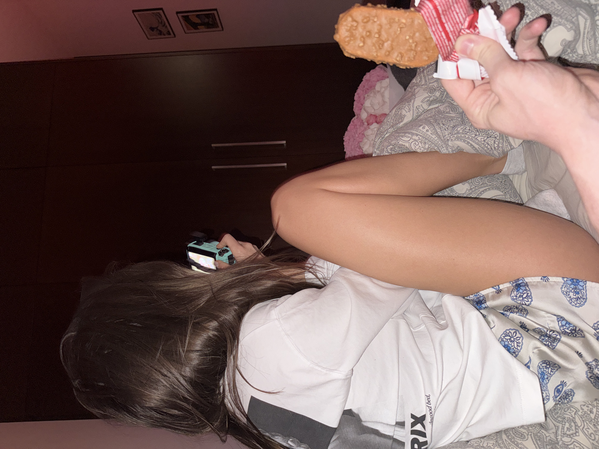 A person is lying in bed in a room with dark wooden furniture. The person is holding a turquoise and black object in their left hand and has long brown hair. A hand in the upper right of the frame is holding a partially unwrapped candy bar over the person. The bed is covered with patterned sheets and a pink blanket. Two framed pictures are on the wall above the furniture. It is not possible to determine if the image was taken in Warsaw, Poland from the given information.