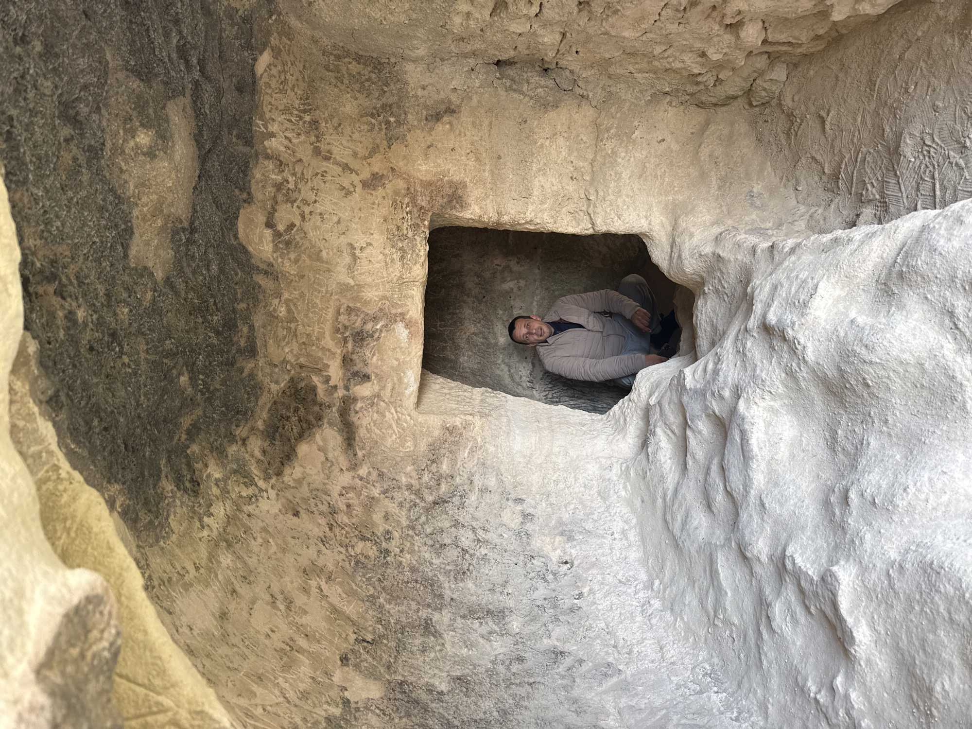 A man is inside a square carved-out space within a larger cave. The cave walls are made of tan-colored rock. The location is in Varna, Bulgaria. The man is looking at the camera. He is wearing a jacket and pants. The space is small, barely large enough for him to sit. The walls of the cave are rough and uneven. There are carvings in the walls.