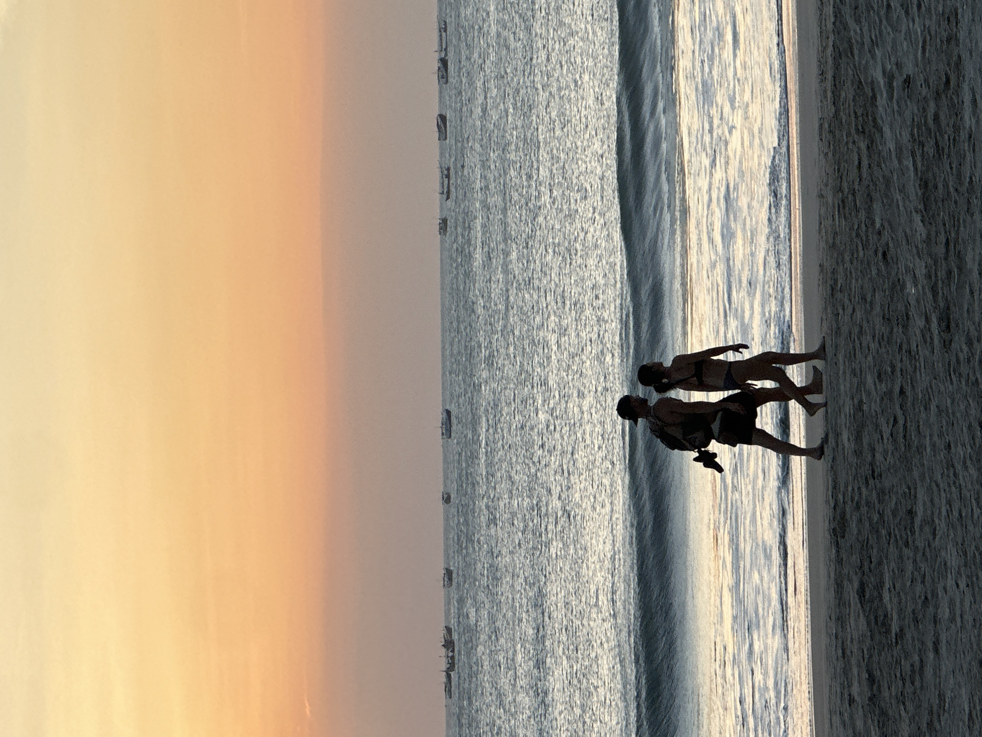 In the city of Kuta, Indonesia, at dusk, a man and a woman walk along a sandy beach. The sky is a gradient of warm colors, transitioning from a pale yellow at the top to a soft orange and pink near the horizon. The sea is calm with gentle waves, reflecting the colors of the sky. Several boats are visible in the distance on the ocean. The couple, silhouetted against the twilight, walks side-by-side towards the left of the frame. The man, wearing a backpack, carries a pair of shoes in his hand. The woman is wearing a bikini. They are walking on the wet sand near the water's edge. The foreground is the beach sand, with the ocean and sky filling the background.