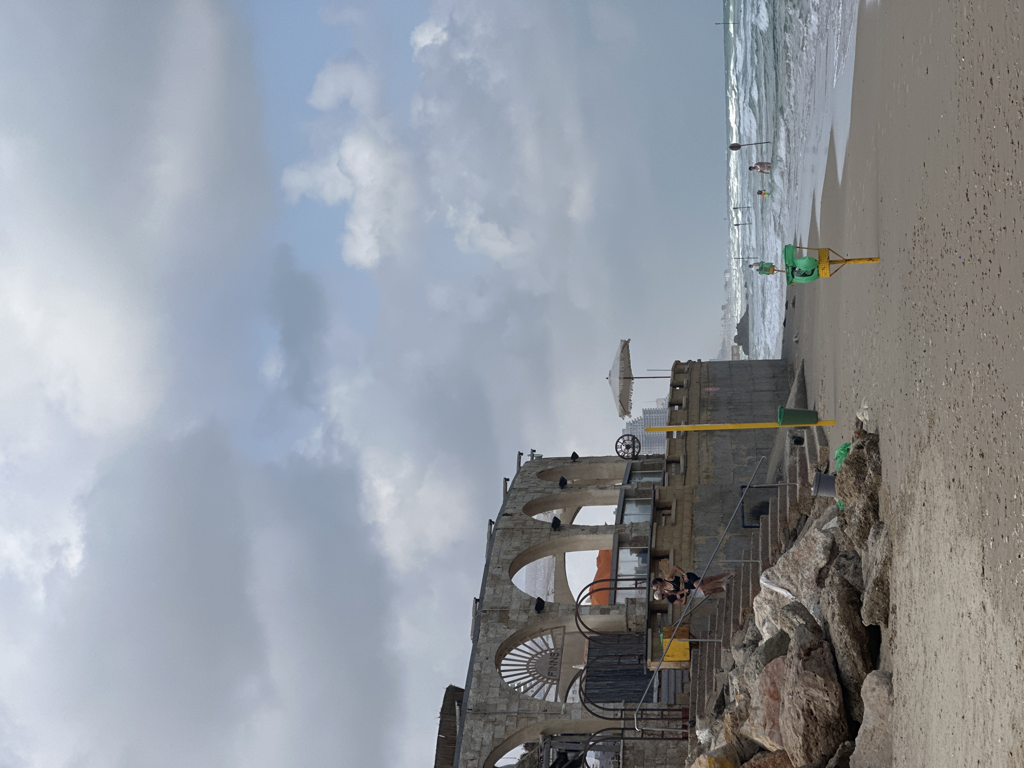 The image depicts a cloudy day at the beach in Netanya, Israel, likely near the Poleg Beach area. A stone building with arched openings and a thatched roof overlooks the sandy beach. Two women are visible on a set of concrete stairs leading up to the building. The sea is choppy with waves, and several people are in the water. Beach equipment, including a white umbrella and a yellow pole with a green bin, are present on the sand. Rocky formations are visible along the shoreline.
