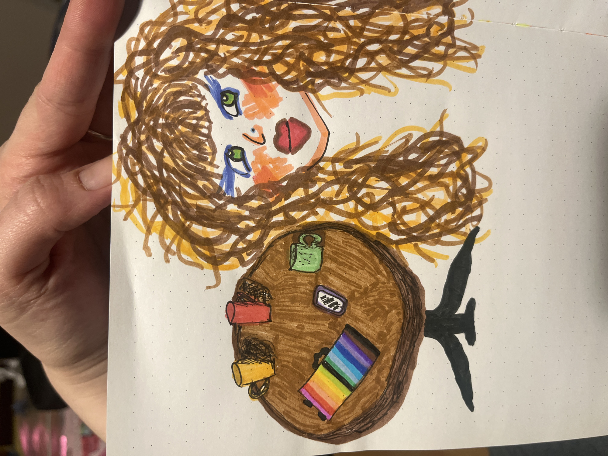 A color marker drawing on a dot grid paper depicts a face at the top with brown and yellow curly hair. The face has green eyes, blue markings around the eyes, and red lips. Below the face is a brown circle representing a table, upon which several objects are placed. A small green box with a black padlock, a gray-tone rectangle, a rainbow-striped rectangle, a red cylinder, and a yellow cylinder are all visible on the table. A black T-shaped element extends from the bottom of the table. The drawing is held by a hand in an indoor setting. The location is not specified.