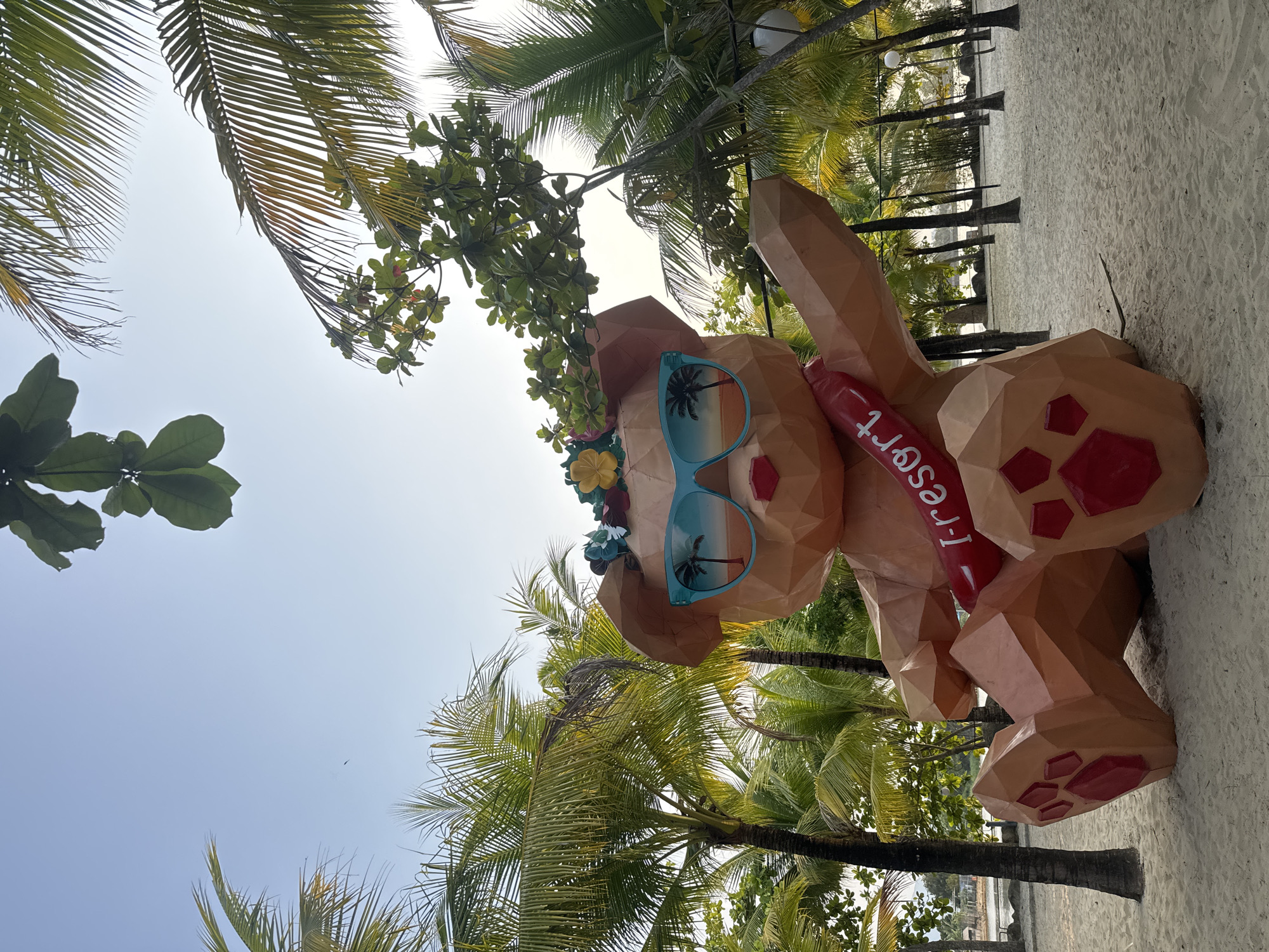 A low-angle shot shows a large, geometrically shaped sculpture of a bear wearing sunglasses and a flower crown, with a red tube labeled "I-Resort," placed on a sandy beach with palm trees. The sky is clear and visible between the trees and branches. The sculpture is the main subject and appears to be an advertisement or landmark for the "I-Resort." The exact location in the city and country is unknown.