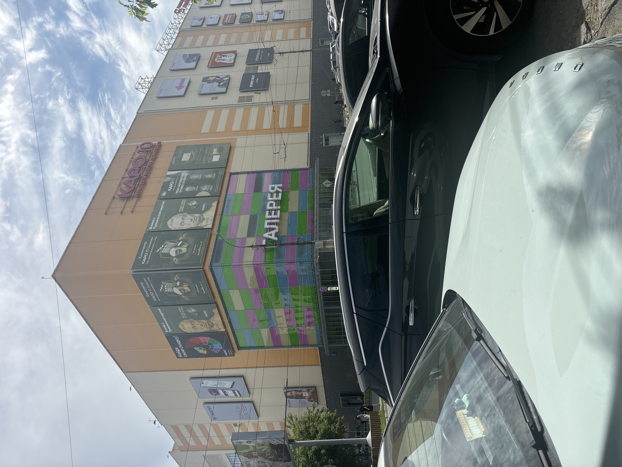 The image depicts a bright, sunny day in front of a large shopping center. The shopping center, named "KARO 10", is a multi-story building with a predominantly orange facade. On the upper levels, there are numerous advertisements and signage. A prominent feature is the colorful, tiled glass facade on the lower levels, displaying the word "ГАЛЕРЕЯ" (Gallery) in large, stylized letters.
In the foreground, two cars are partially visible, parked. The closest car is light-colored, with its hood and windshield in view. Behind it, a dark-colored SUV is parked. A wooden fence and some vegetation are visible to the left of the shopping center entrance. A few pedestrians can be seen walking near the entrance of the shopping center. The sky is partly cloudy with patches of blue. The presence of "KARO 10" signage and the Cyrillic text on the building strongly suggests the location is in Russia. Based on external information, "KARO 10" is a cinema and shopping complex located in Novosibirsk, Russia.
