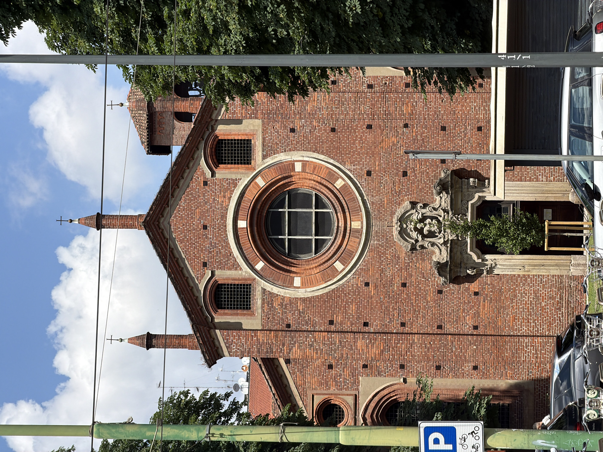 The image, oriented 90 degrees clockwise, displays the detailed facade of a brick building, characteristic of a church or historical structure, set against a bright, partly cloudy daytime sky. The architecture prominently features reddish-brown bricks, with lighter-colored stone accents around the windows. A large circular window, a rose window, with a grid pattern, dominates the central part of the facade. Smaller arched windows with metal grates are visible on either side of the main facade and below the rose window.
Two slender spires, each topped with a cross, rise from the roofline, which is covered with terracotta tiles. To the right of the main structure, an elaborate stone carving, possibly a fountain or decorative element, adds architectural detail. Green foliage from trees is visible at the top and bottom of the frame, suggesting a treelined street. Utility poles and wires traverse the foreground.
In the lower right, parked vehicles, including parts of a car and several bicycles, are visible. A blue parking sign with a white "P" and a bicycle icon is clearly depicted. The text "41/1" is visible on a grey pole. The scene is static, capturing an architectural view, likely in Milan, Italy, under natural daylight.