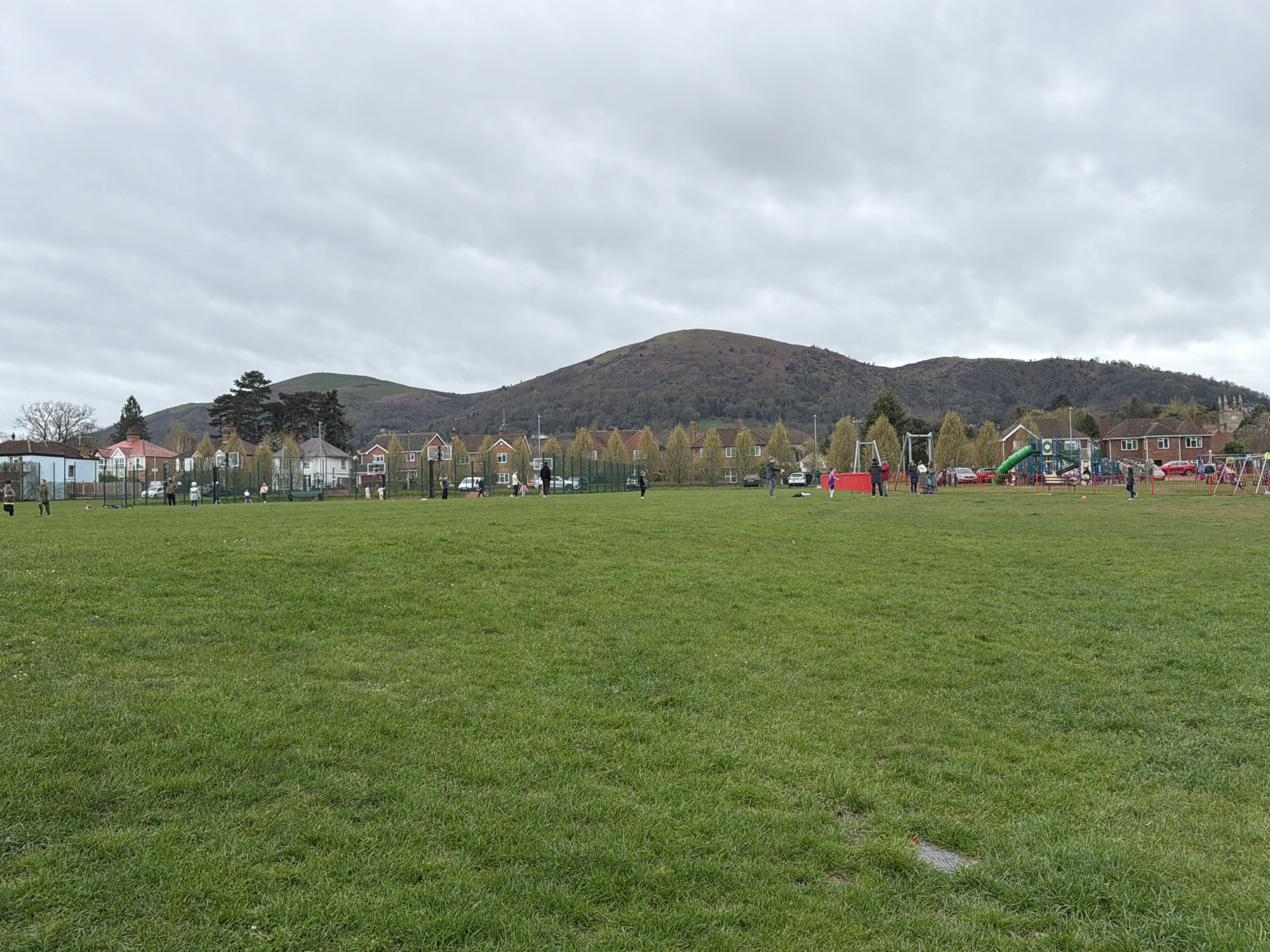 The image depicts a large grassy field in the Malvern Hills, United Kingdom. In the background, a mountain rises behind houses and trees. A playground is visible to the right. Several people are scattered across the field, some playing games and others standing in groups. The sky is overcast.