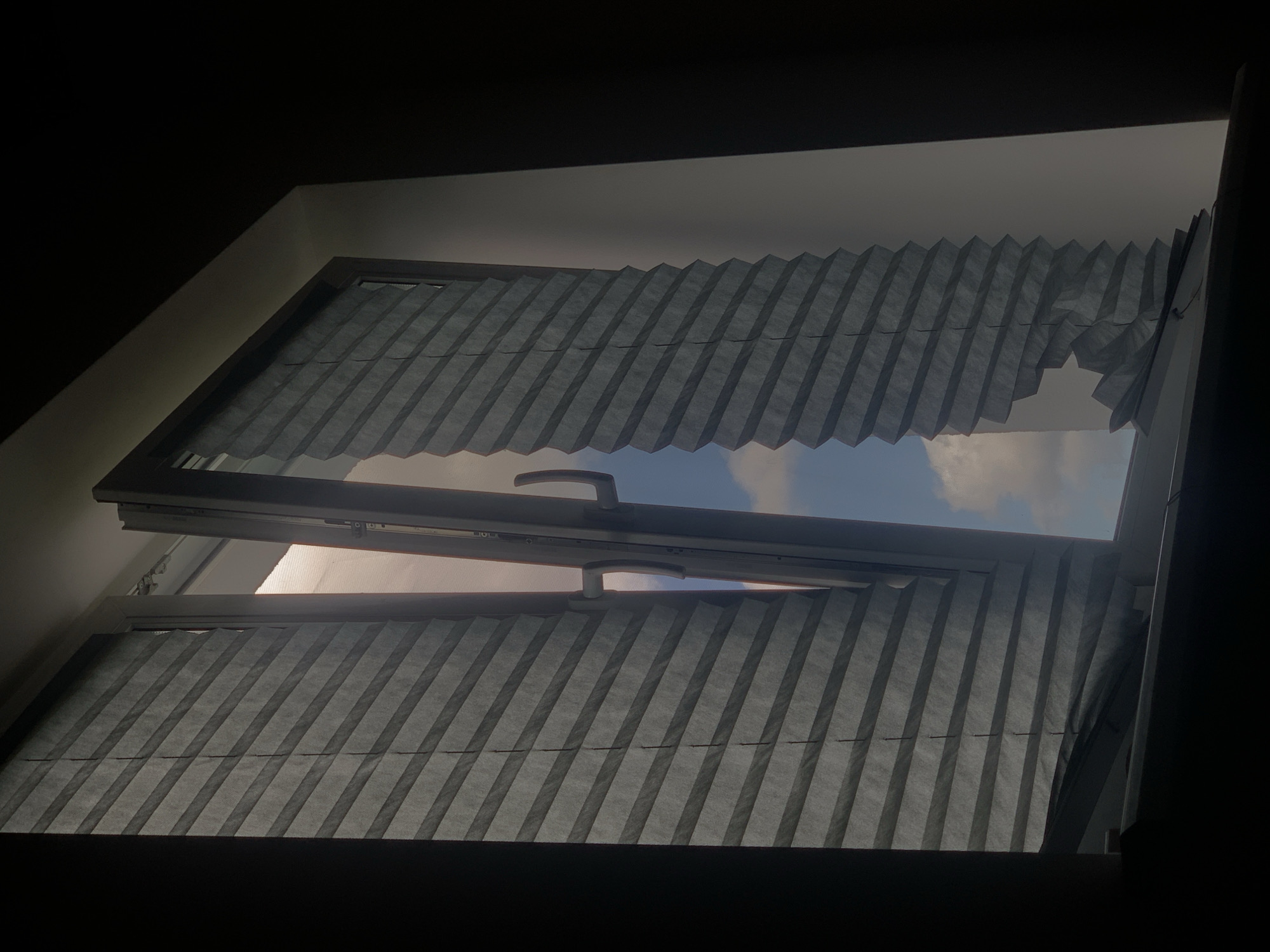 The image shows an interior view of an open window in a dimly lit room. The window frame is white, and the window itself is open outwards. On either side of the window, there are pleated blinds, dark grey in color. The blinds on the left are mostly intact, but the blind on the right appears to be torn or damaged at the bottom. Through the open window, a bright, partly cloudy sky is visible, suggesting it is daytime. The scene is consistent with an apartment or residential building. The location is described as Minsk, Belarus, though no specific landmarks or indicators of this location are present within the image itself.