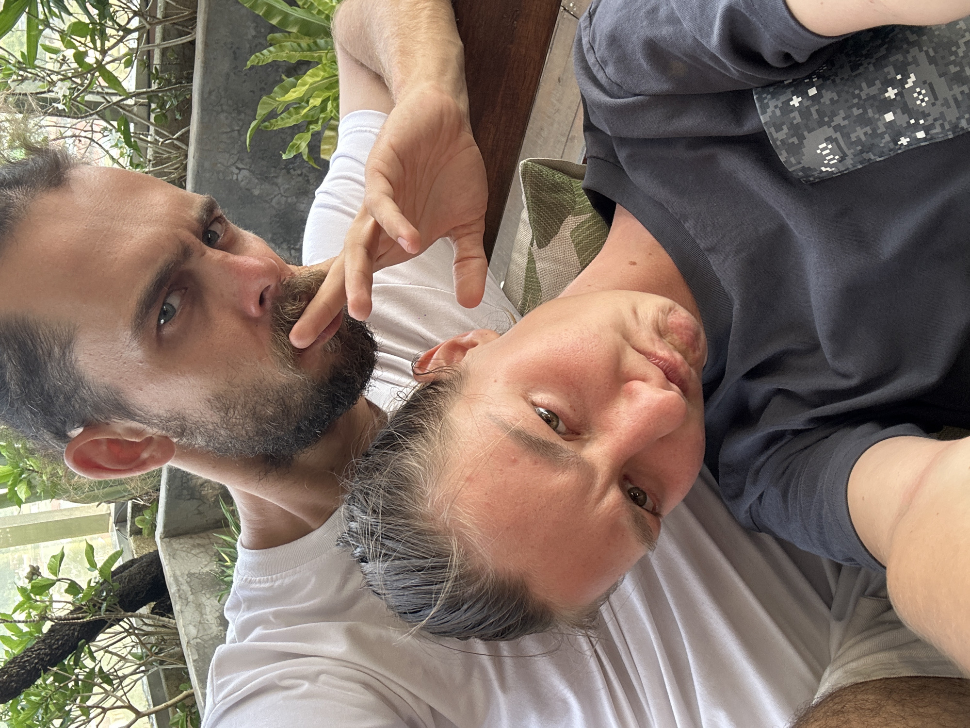 The image is a close-up, angled selfie-style portrait of two individuals, a man and a woman, in what appears to be an outdoor or semi-outdoor setting.
On the left, a man with a dark beard and mustache looks directly into the camera with an intense gaze from his light-colored eyes. He is wearing a plain white t-shirt, and his right hand is raised with fingers near his chin and mouth.
Adjacent to him, on the right, a woman also looks directly into the camera. She has fair skin, rosy cheeks, and light brown eyes, with reddish-blonde hair showing visible grey strands. She is wearing a dark grey t-shirt that features a grey and white digital camouflage patterned pocket on the chest. Her expression is relaxed and engaging.
The background shows lush green tropical foliage and hints of a concrete structure, suggesting a garden, patio, or balcony setting. A wooden surface is visible between the two, and a green and white checkered cushion is partially seen behind the woman. The lighting suggests it is daytime. Both individuals appear to be posing casually for the photograph. There is no visible text in the image.