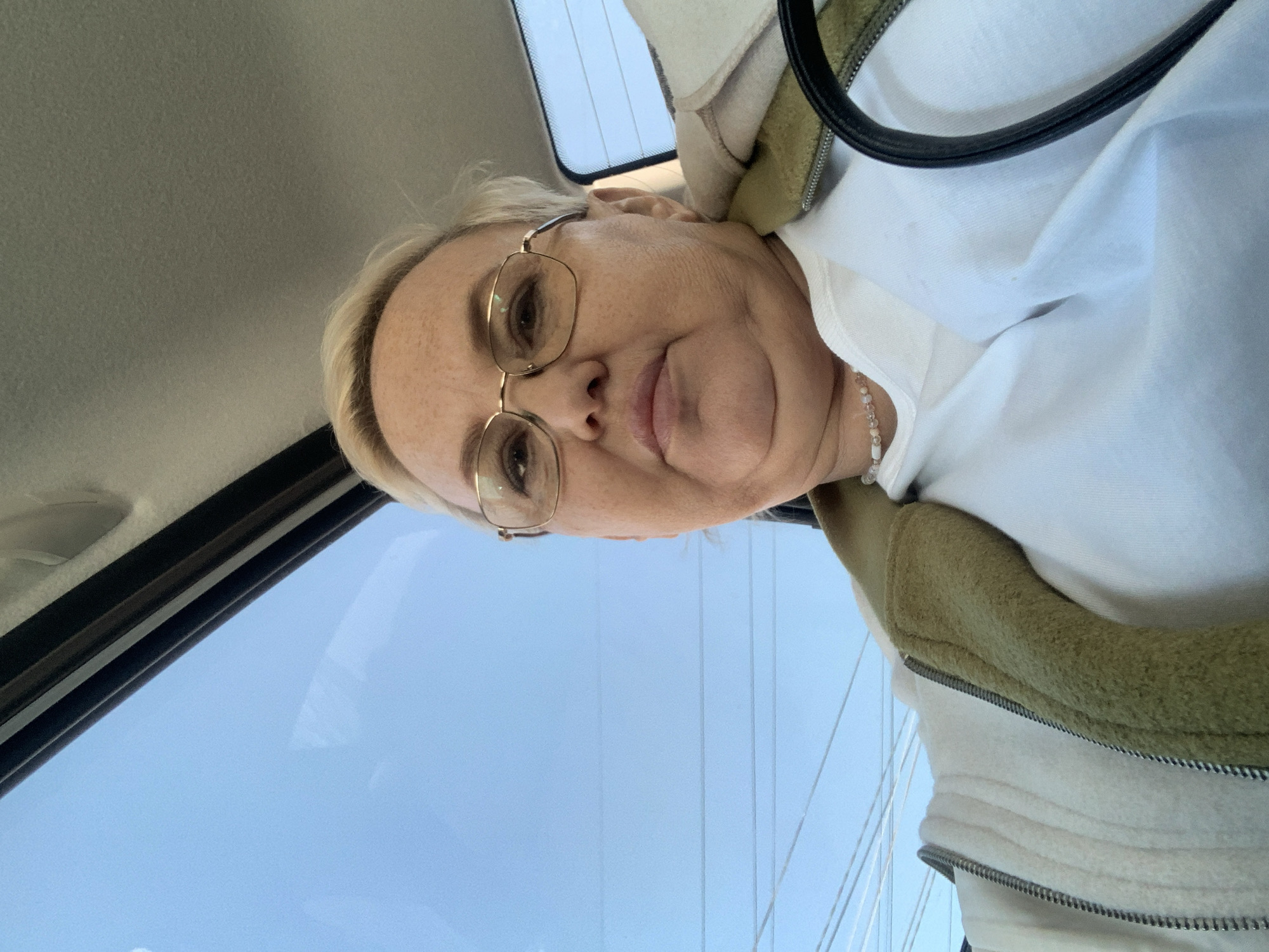 The image shows a selfie of a woman wearing glasses, a white shirt, and a jacket with a green fleece lining. She is likely in a car, as the interior of the vehicle and a glimpse of the sky and overhead power lines are visible in the background. It is not possible to confirm her location in the city of Chaikovsky, Russia, from this image alone.