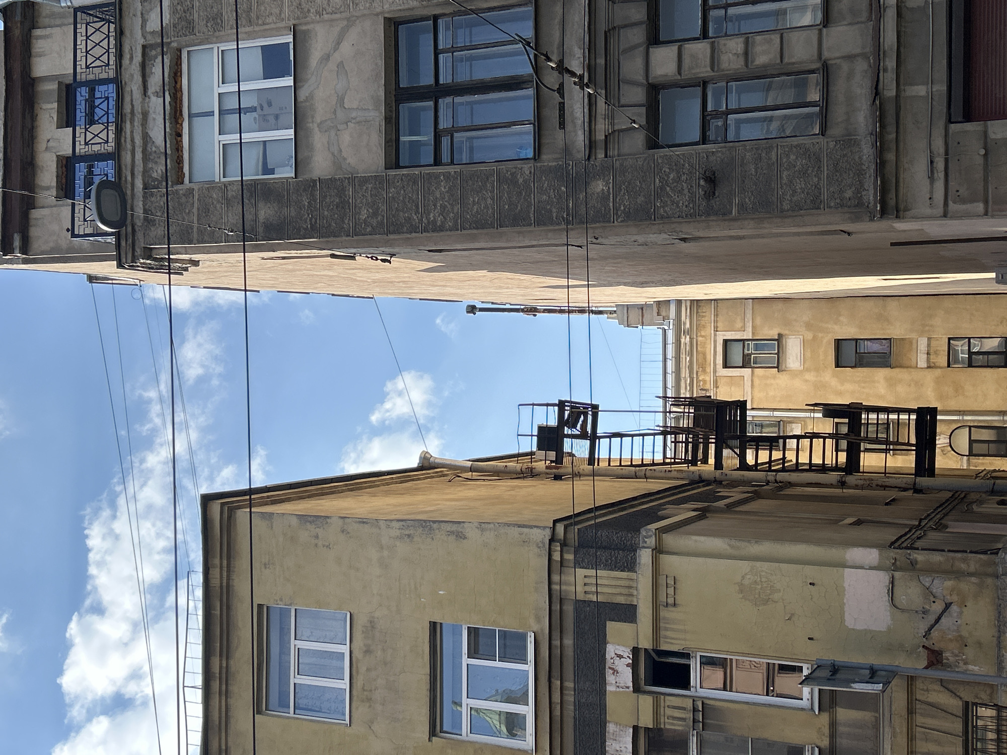 An outdoor, medium shot captures the side of two buildings in an urban environment, under a blue sky with scattered clouds. The building on the left is painted a faded yellow, with several windows visible. The building on the right is a weathered, grey structure. A black metal fire escape is attached to the yellow building, with multiple levels and ladders. Power lines are strung across the sky in front of both buildings. The lower portion of the image shows the base of the grey building, with a window and what appears to be a red shuttered opening. The yellow building has a downspout running along its exterior and some signs of wear and tear on its facade. The overall impression is one of an older, somewhat neglected part of a city.