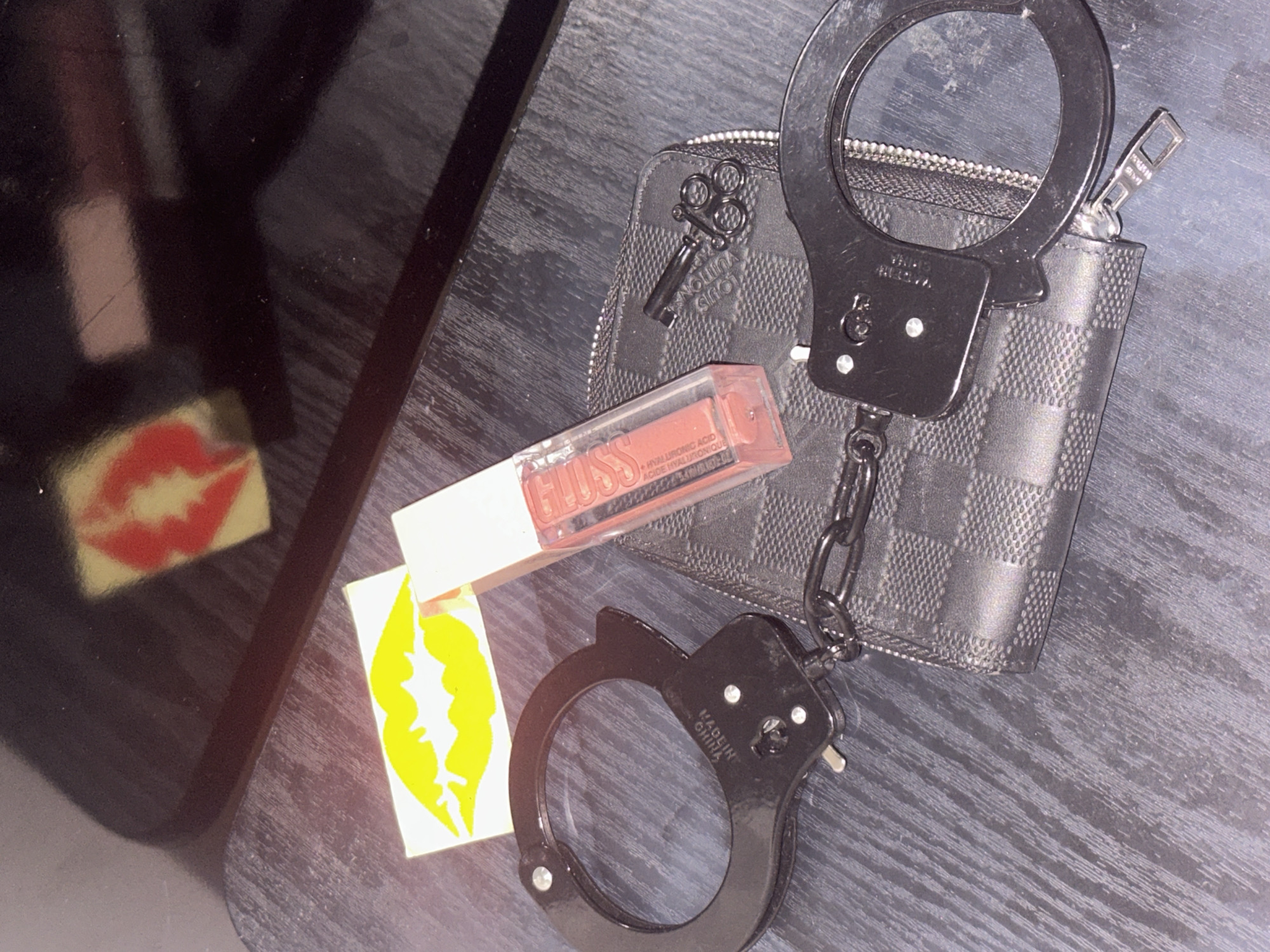 The scene depicts an indoor setting where several personal items are arranged on a dark, textured tabletop. A pair of black metal handcuffs, linked by a short chain, is centrally positioned. One cuff is visibly stamped with "MADE IN CHINA." A small, black, stylized key, accompanied by three small black rings, is attached to the handcuff's chain. Adjacent to the handcuffs is a small, zippered, black wallet or pouch, exhibiting a dark grey and black checkered pattern, with the text "LOUIS VUITTON" visible in an inverted orientation. A clear, rectangular tube of "GUESS GLOSS" lip gloss, containing a peachy-nude liquid and marked with "HYALURONIC ACID," rests near the wallet. A white rectangular sticker featuring a bright yellow lip silhouette is placed directly on the tabletop. In the upper background, a dark, reflective surface, possibly a screen or polished furniture, shows a blurred reflection of a red lip silhouette on a yellow background. The tabletop surface appears to be dark wood or laminate with a visible linear grain. The lighting suggests an artificial source, creating reflections on the glossy items and surfaces. The image contains no discernible visual markers, such as landmarks or street signs, to identify a specific location within Sofia, Bulgaria.