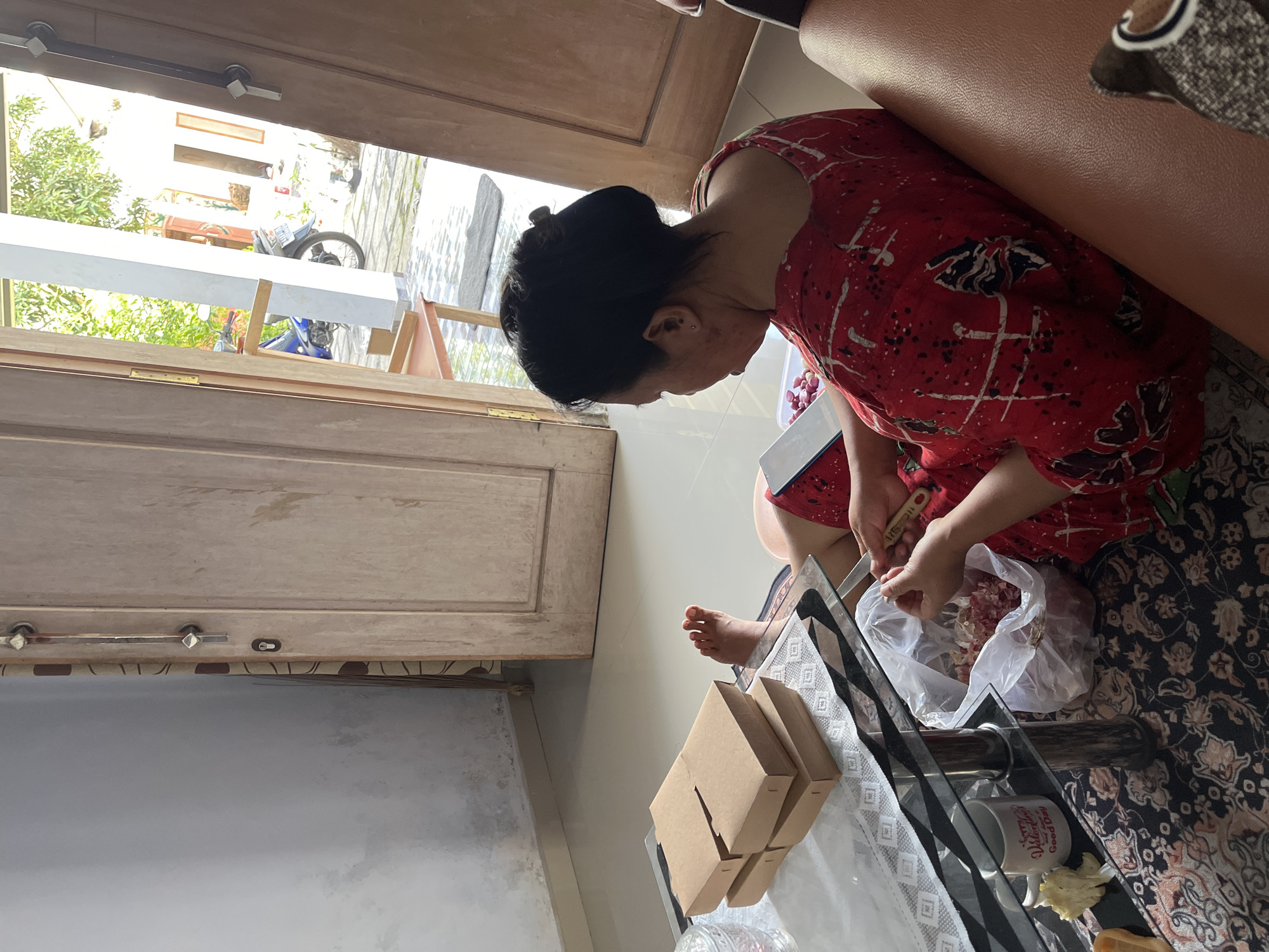 The image depicts an interior scene likely within a residential dwelling in Jambearum, Indonesia. The focus is on a woman, viewed from behind and to her side, wearing a red and white patterned garment. She is seated on the floor, leaning towards a glass-topped coffee table. Her hands are engaged in an activity, appearing to be preparing food, with a plastic bag containing diced meat and a knife visible. The table also holds stacked brown cardboard boxes and a white mug. In the background, a wooden door is ajar, revealing a sunlit outdoor area with a motorcycle and foliage. The floor is tiled, and a patterned rug is visible beneath the table and the woman. The overall impression is one of domestic activity within a home.