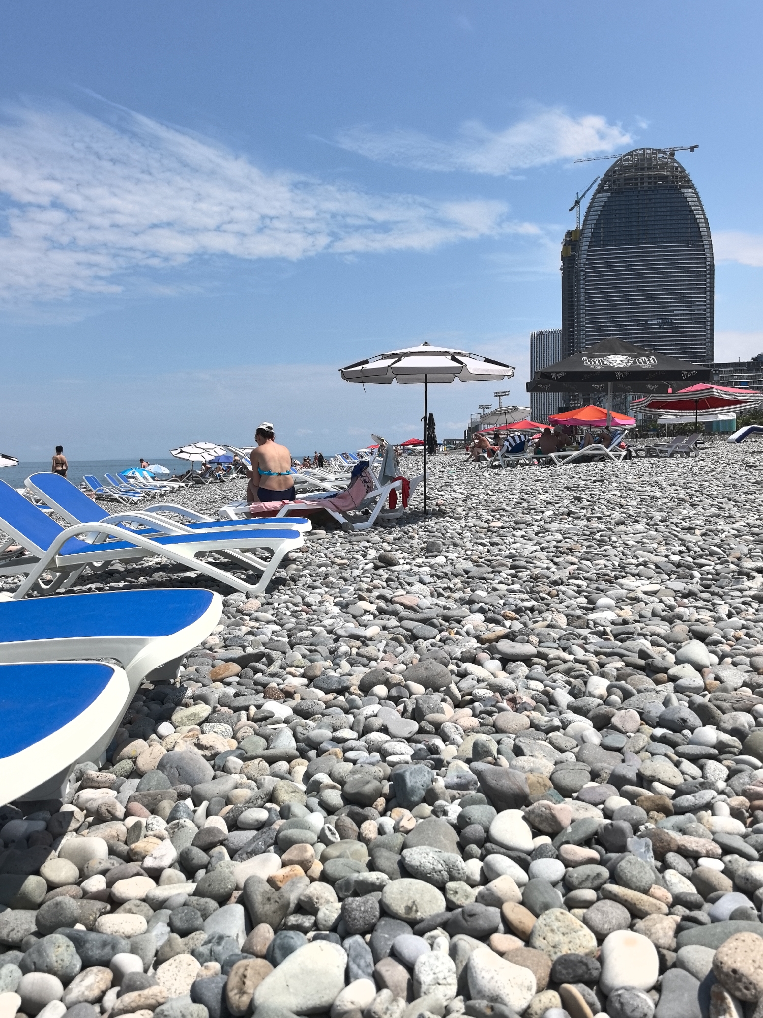 The image shows a pebble beach with many people relaxing on lounge chairs under umbrellas. In the background, a modern skyscraper is under construction. The sky is blue with scattered white clouds. The overall scene depicts a popular tourist destination on a sunny day.