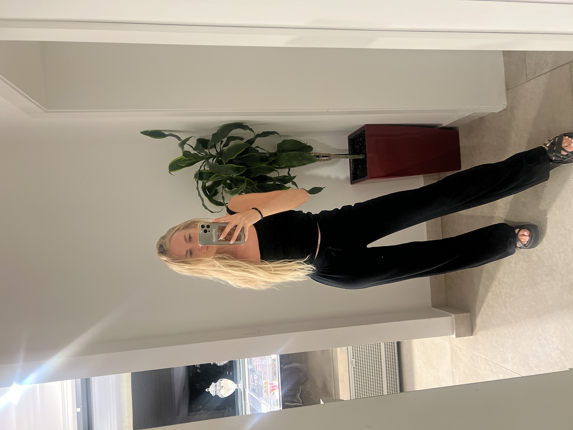 A young Caucasian woman with long blonde hair is taking a mirror selfie indoors. She is wearing a black strapless top and black wide-leg pants. She has on black sandals with gold buckles. She is holding a smartphone in her right hand, and a black hair tie is on her left wrist. Behind her, to her right, is a tall green plant in a square red planter. To her left, a display case containing various sweets is visible. The floor is tiled with a light beige. The wall behind her is white. The scene appears to be in a commercial establishment, possibly a sweet shop or cafe, located in Boreti, Montenegro.