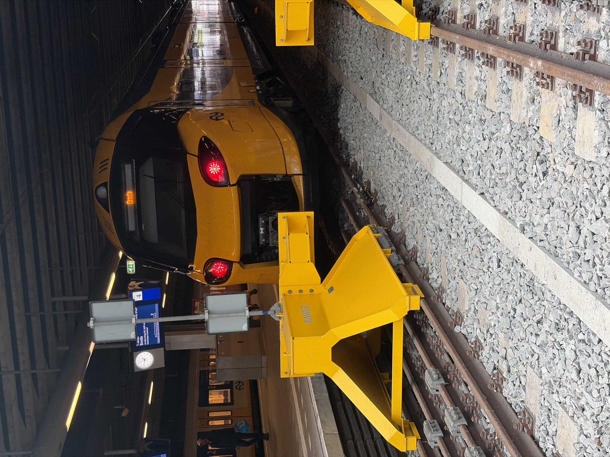 The image captures a modern yellow and grey train, likely a Dutch Sprinter model, at a railway station platform. The perspective is from the side, looking towards the front of the train, which is currently at the end of the track. The train has two prominent red taillights illuminated on its rear/front (depending on direction of travel) and a digital destination display that appears to show "SPRINTER" in orange text. The number "22" is visible as a logo on the side of the train's front carriage.
In the immediate foreground, a bright yellow railway buffer stop or derailer is prominently positioned on the tracks, indicating the end of the line. Another similar yellow structure is visible further down the track. The word "HAWKIE" is faintly visible on the side of the closer yellow structure. The tracks are laid on a bed of grey ballast with concrete sleepers.
The station environment includes a dark, covered overhead structure, suggesting an indoor or sheltered platform. Artificial lighting strips run along the ceiling. On the platform, a grey pole holds an analog clock, showing approximately 1:00 or 1:05, and a digital information display. This display shows "Breda", "Delft", and "Rotterdam Centraal" as destinations, along with "4" indicating the platform number. A person wearing a blue top is visible on the platform near a train door.
The scene depicts a stationary train at a platform, possibly awaiting departure or having just arrived, at what appears to be a major railway station in the Netherlands, consistent with The Hague.