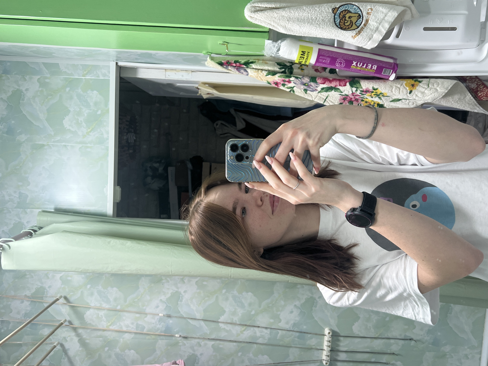 An image depicts a young female taking a mirror selfie inside a bathroom, likely located in Chaikovsky, Russia. The female, with light brown hair and freckles, is wearing a white short-sleeved t-shirt featuring a dark circular graphic with a light blue cartoon character. She holds a light blue patterned smartphone with three rear cameras in her right hand, positioned to capture her reflection. A silver ring is visible on her right ring finger, and her fingernails have a light pink polish. On her left wrist, she wears a black sports watch with a circular face.
The bathroom environment features walls covered in light green and white marble-patterned panels. To the left of the subject, a wall-mounted retractable clothes drying rack with multiple horizontal rods is visible. A light green shower curtain hangs from a rod in the central background. Through an open doorway to the left of the curtain, a dimly lit room with exposed brick-like wall textures and stored items is discernible.
To the right of the subject, a narrow bright green vertical panel is present. Below it, two hooks hold two towels. The towel on the left is white with a floral pattern (pink, yellow, green). The towel on the right is white and features an embroidered ram's head design above Cyrillic text, which clearly spells "ЧАЙКОВСКИЙ" (Chaikovsky), confirming the city location. Adjacent to the towels, a white bottle labeled "DETOX" and "MEI" (visible in reverse due to the mirror) is hanging upside down in a clear plastic bag containing cotton pads. The top portion of a white appliance, consistent with a washing machine or dryer, is partially visible below the towels and cosmetic product.