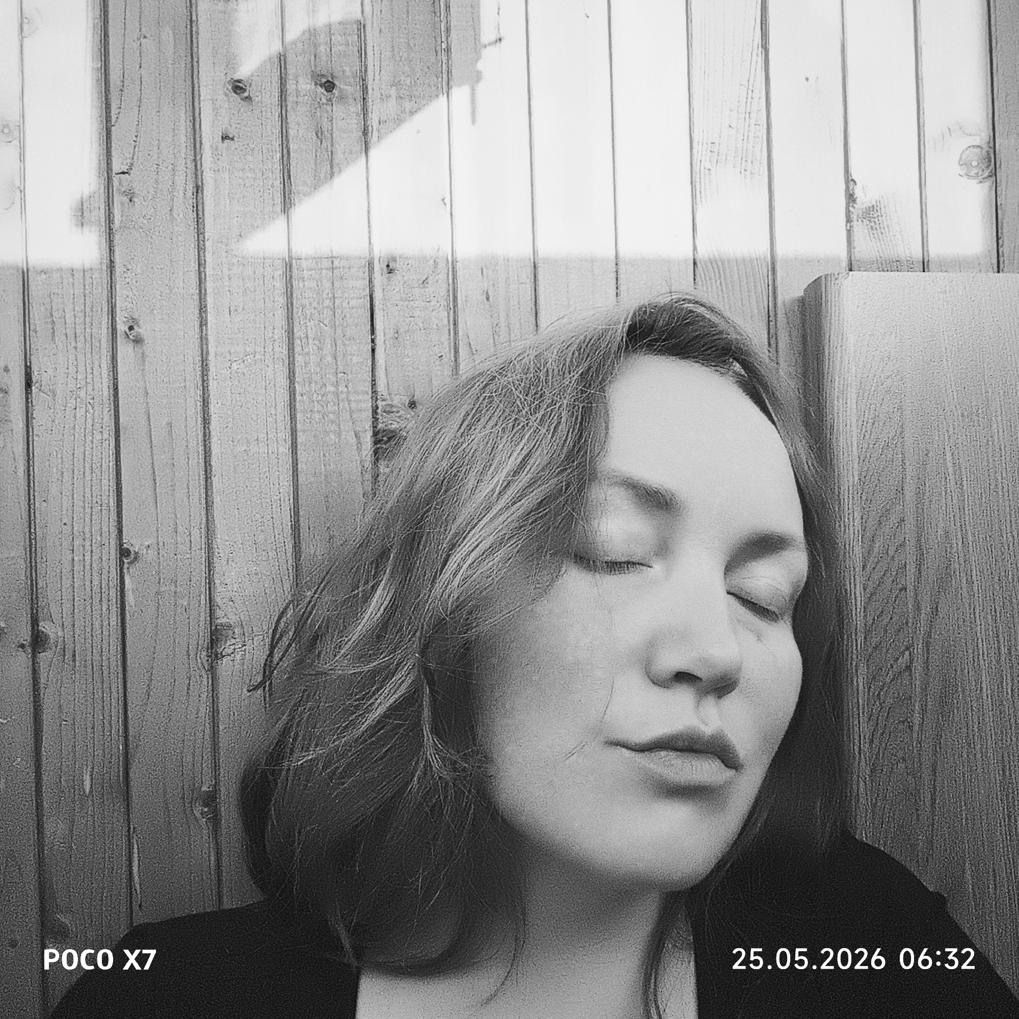 The image is a black and white close-up of a woman with her eyes closed, appearing to be resting or sleeping. Her face is pale, and her hair is shoulder-length and wavy. Behind her is a wooden fence or wall, partially illuminated by sunlight. A shadow of an indeterminate object is cast on the wall. The bottom left corner of the image displays "POCO X7," indicating the device used to take the photograph. In the bottom right corner, "25.05.2026 06:32" is displayed, suggesting the date and time the photo was taken. The location is unspecified, indicated by Unknown, Unknown. There are no visible actions or interactions beyond the woman's state of rest.