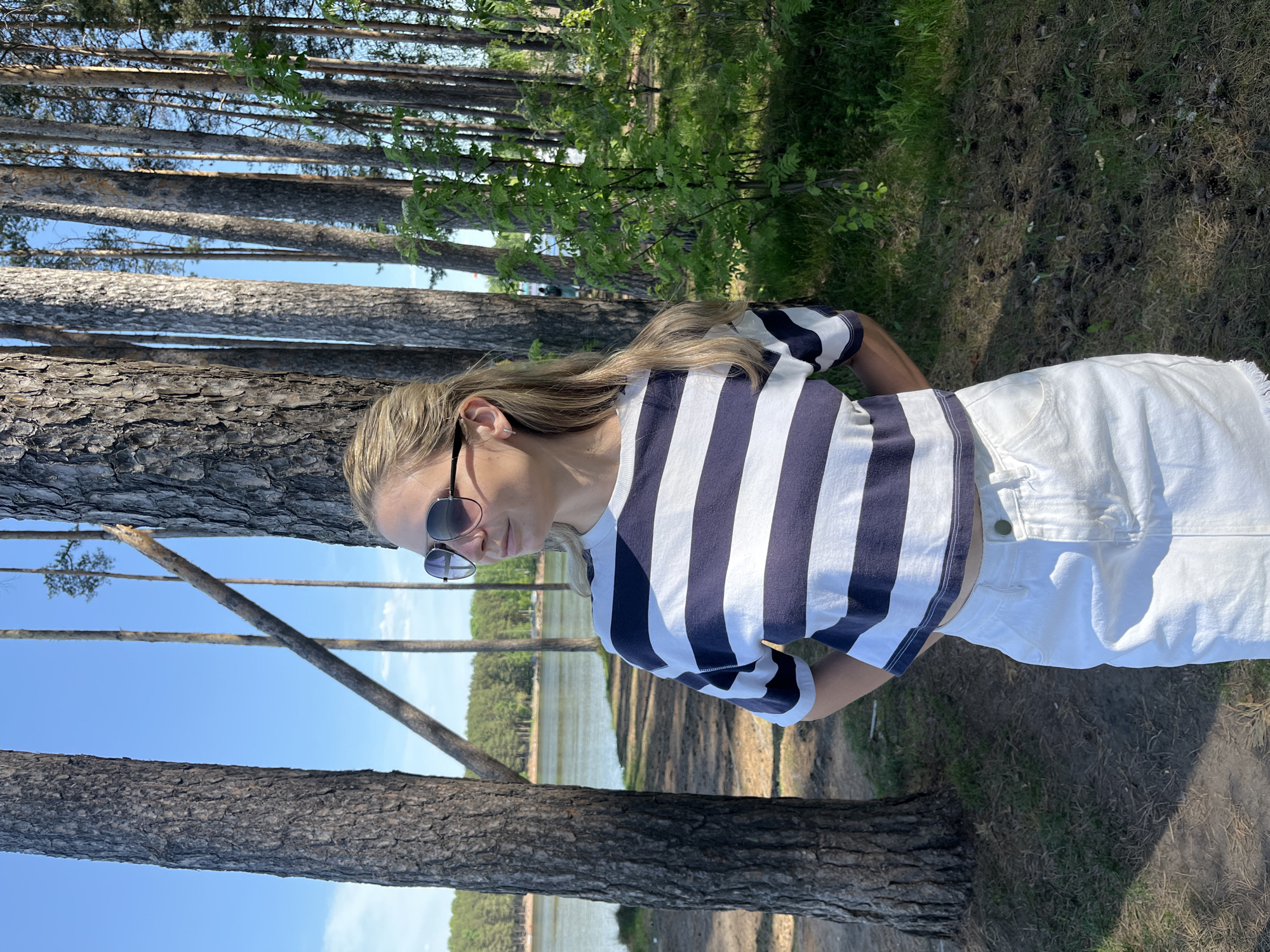 A young woman stands outdoors near a forest in Chaikovsky, Russia. She is wearing sunglasses, a striped shirt, and white shorts. The scene includes tall pine trees in the background and a view of water in the distance. The woman is positioned next to a tree trunk, and the ground is covered with grass and shadows.
