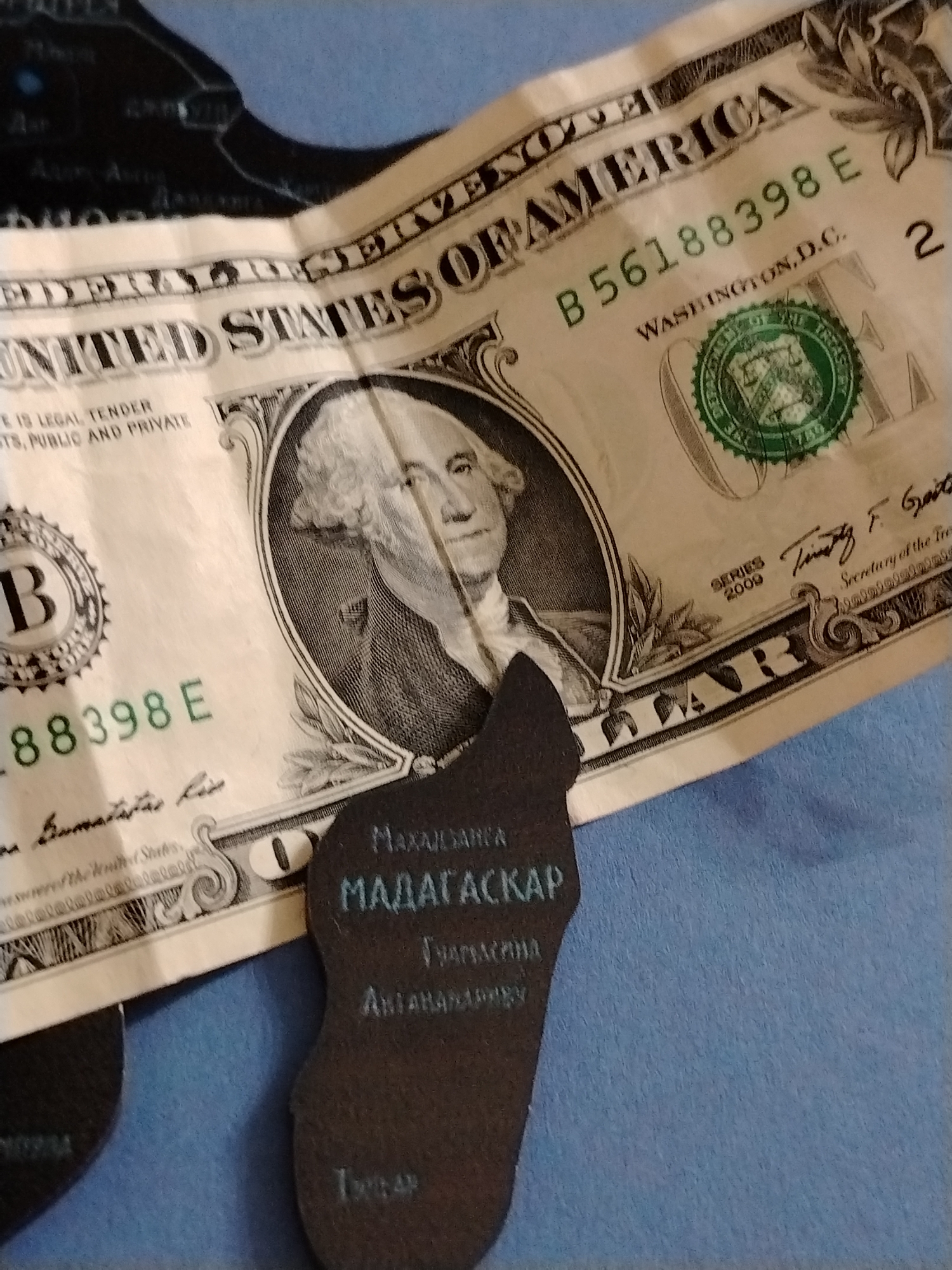 The image is a close-up of a one-dollar bill and a small, dark, irregularly shaped cutout that resembles a map fragment. The dollar bill is wrinkled and shows a portion of George Washington's portrait. Text on the bill is visible, including "UNITED STATES OF AMERICA", a serial number starting with "B 56188398 E", and "WASHINGTON, D.C.". A circular seal with an eagle is also visible.
The cutout has text printed on it in a lighter color. The most prominent text reads "МАДАГАСКАР" (Madagascar). Below this are other words that appear to be place names, possibly cities or regions, in what appears to be Cyrillic script. One line reads "ГУАРАСИМА" and another "АНТАМАНАРУ". At the very bottom of the cutout, a single word "ТУАР" is visible.
The objects are placed on a blue surface, possibly fabric or paper. The background is out of focus. There are no people visible in the image. The lighting appears to be from above and slightly to the left.
There is no information in the image that indicates a location in Krasnodar, Russia. The text on the cutout refers to Madagascar, an island country off the southeast coast of Africa.