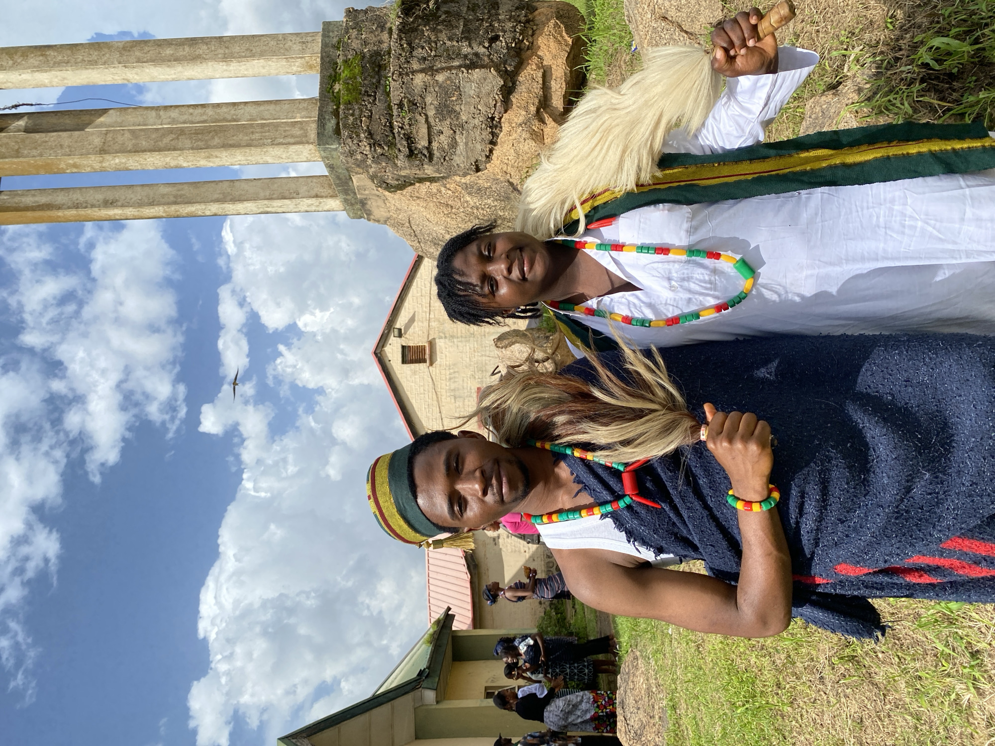 Two individuals, adorned in traditional attire, are captured in a low-angle shot against a backdrop of a cloudy blue sky and lush green grass. The person on the left, wearing a white garment with a green and gold trim, has a white, furry accessory around their neck and is holding a wooden object. Their hair is dark, and they are smiling. The person on the right wears a dark blue garment over a white shirt, with a beaded necklace and bracelet in red, yellow, and green. They are also wearing a colorful cap with a tassel. Both individuals appear to be of African descent.
The setting includes a light-colored building with a red roof and several concrete structures in the background. A bird is visible flying in the sky. The overall atmosphere suggests an outdoor event or gathering, possibly a cultural ceremony or celebration, given the traditional clothing. The lighting indicates it is daytime, with the sun casting shadows, and the presence of clouds suggests variable weather conditions. There is no visible text in the image. The emotions conveyed are positive, with smiles and a relaxed posture. The location context is identified as Wang, Nigeria.