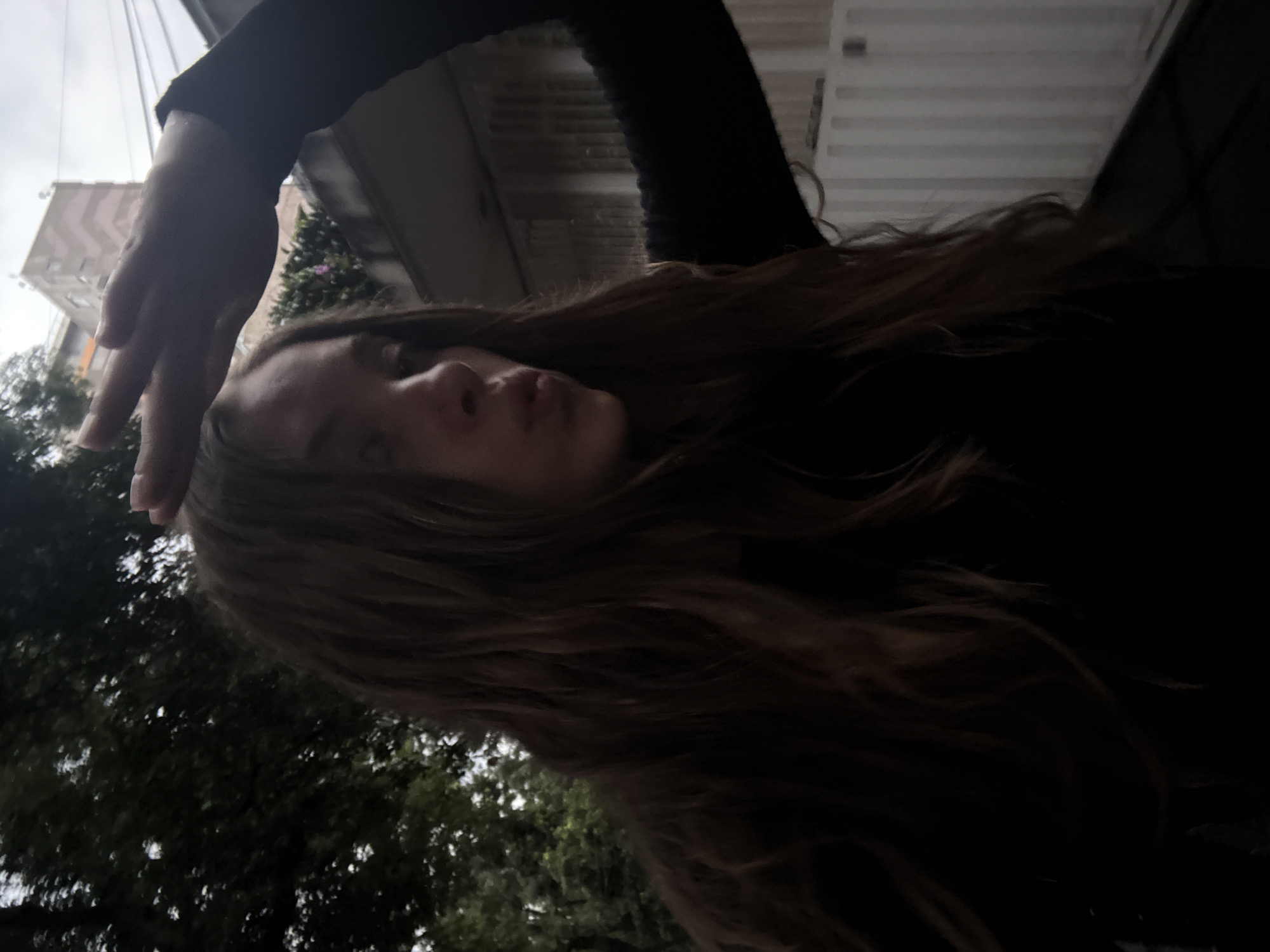 A young woman with long, wavy brown hair is in the foreground, holding her right hand up near her forehead, obscuring part of her face and creating a shadow. Her lips are pursed. She is wearing a black long-sleeved shirt. Behind her, to the left, are dark green trees. To the right, there is a light-colored building with a grid-like facade, and a taller building in the background, suggesting an urban environment. The sky is overcast. The overall lighting is dim. The scene appears to be outdoors, possibly in a residential or park area within Mexico City, Mexico, given the architectural styles of the buildings and the presence of lush vegetation.